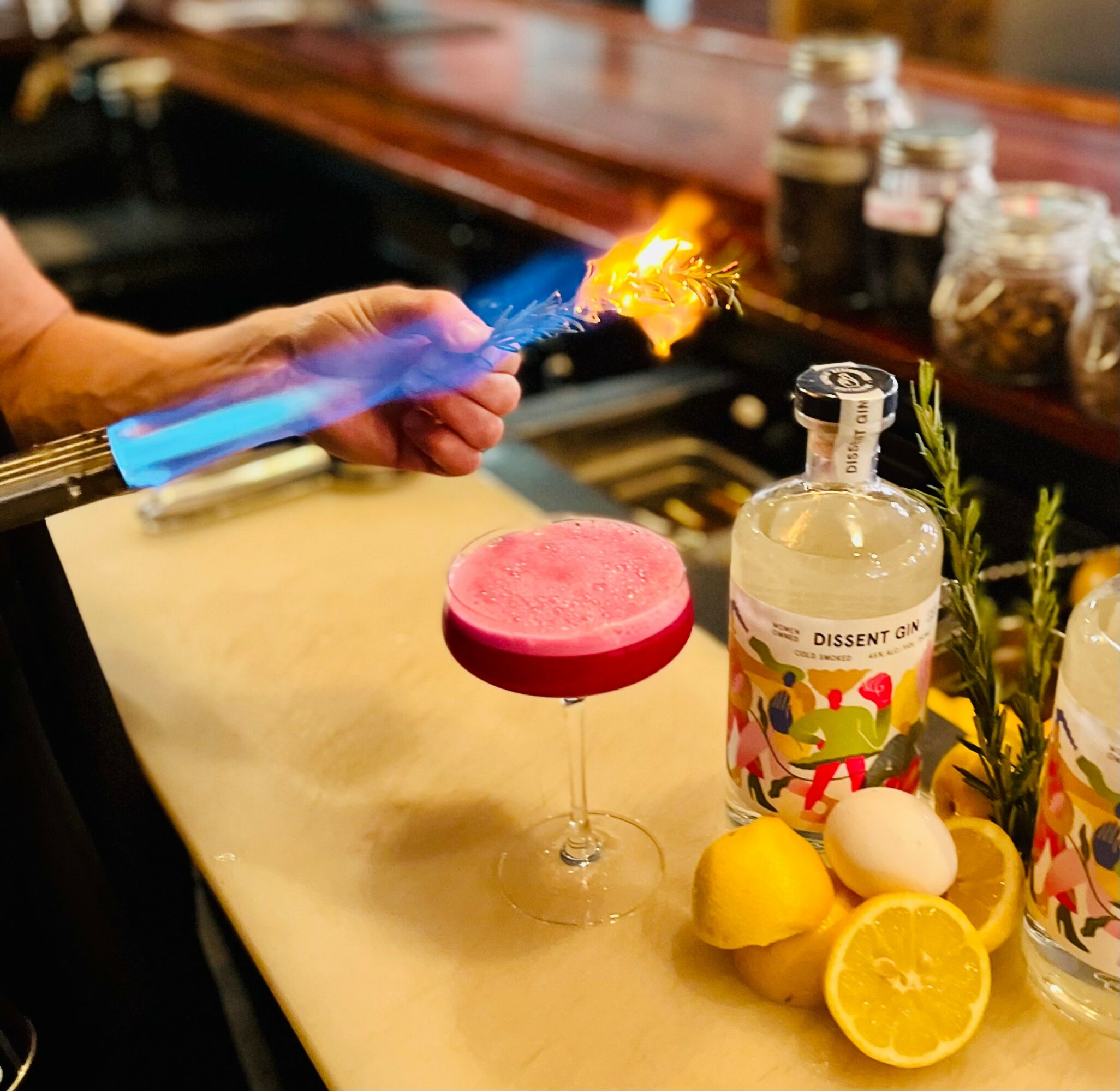 Person holding a flame over a pink cocktail glass with lemons, eggs, and herbs nearby, bar counter in background.