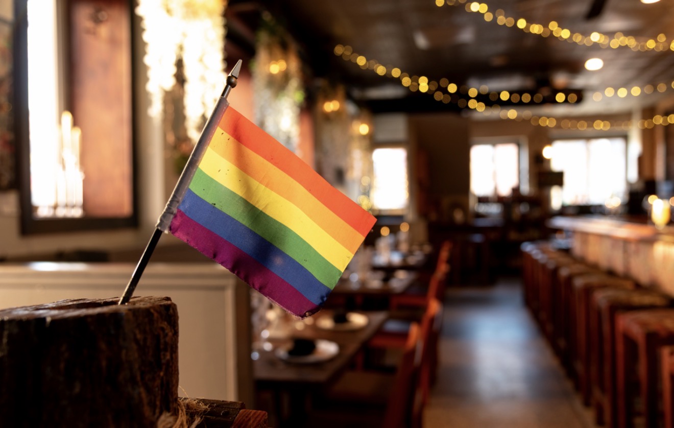 Rainbow pride flag on a table in a warmly lit restaurant with string lights and wooden furniture.