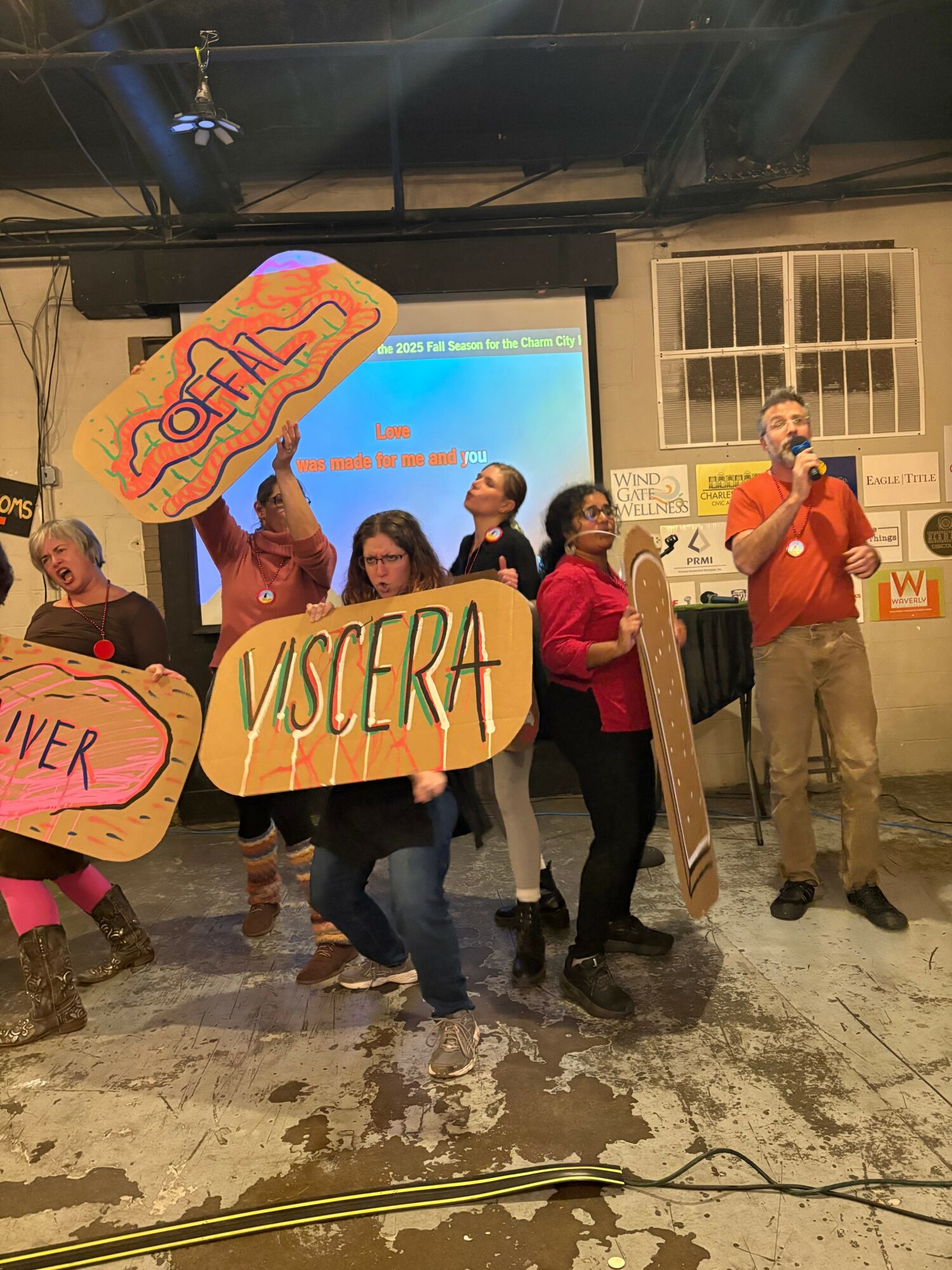 Group of people holding colorful signs, one reading 'VISCERA,' in an indoor setting with a speaker and window behind them.