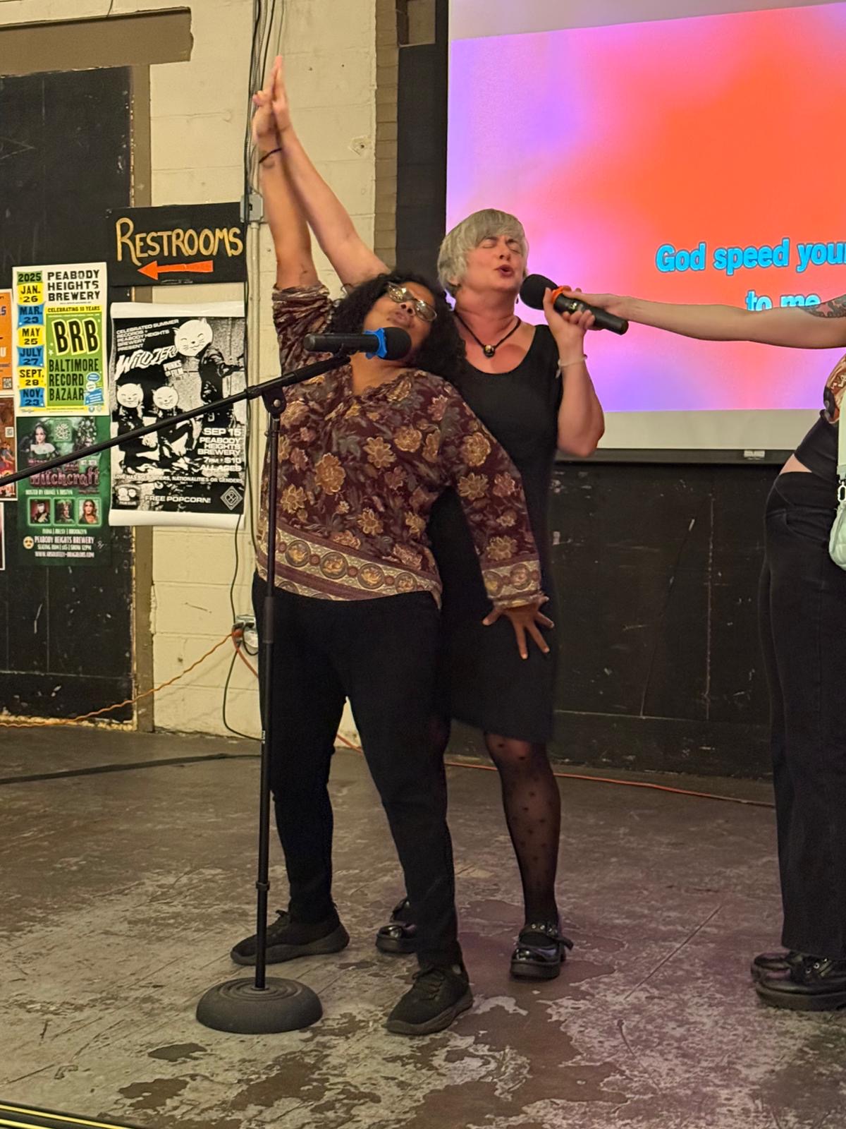 Two women stand on stage, one holding a microphone, with a large screen behind them displaying colorful text. One woman raises her hand.
