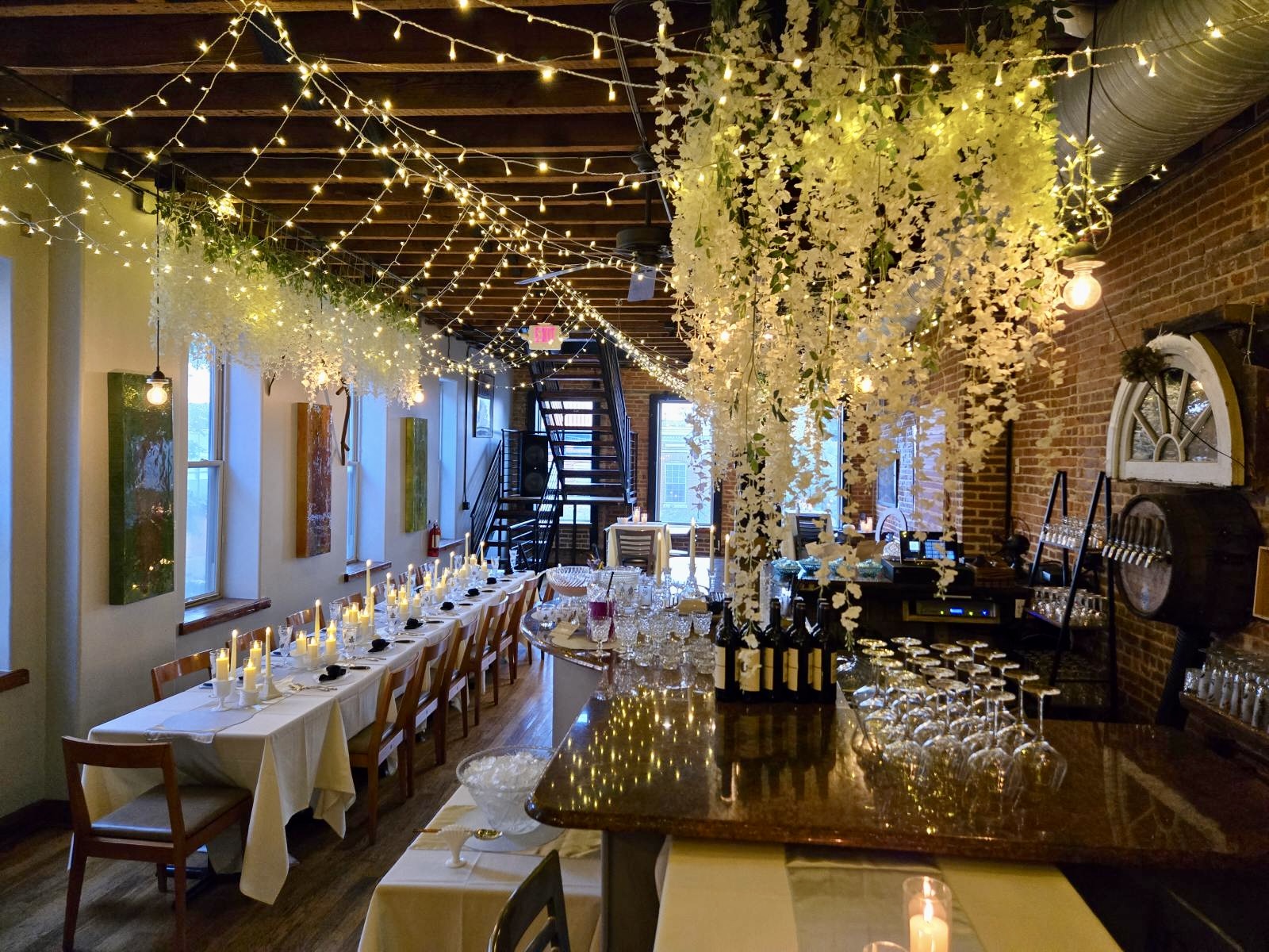 Long dining table with candles, flowers, and glasses, decorated with string lights and hanging flowers in a warmly lit room.