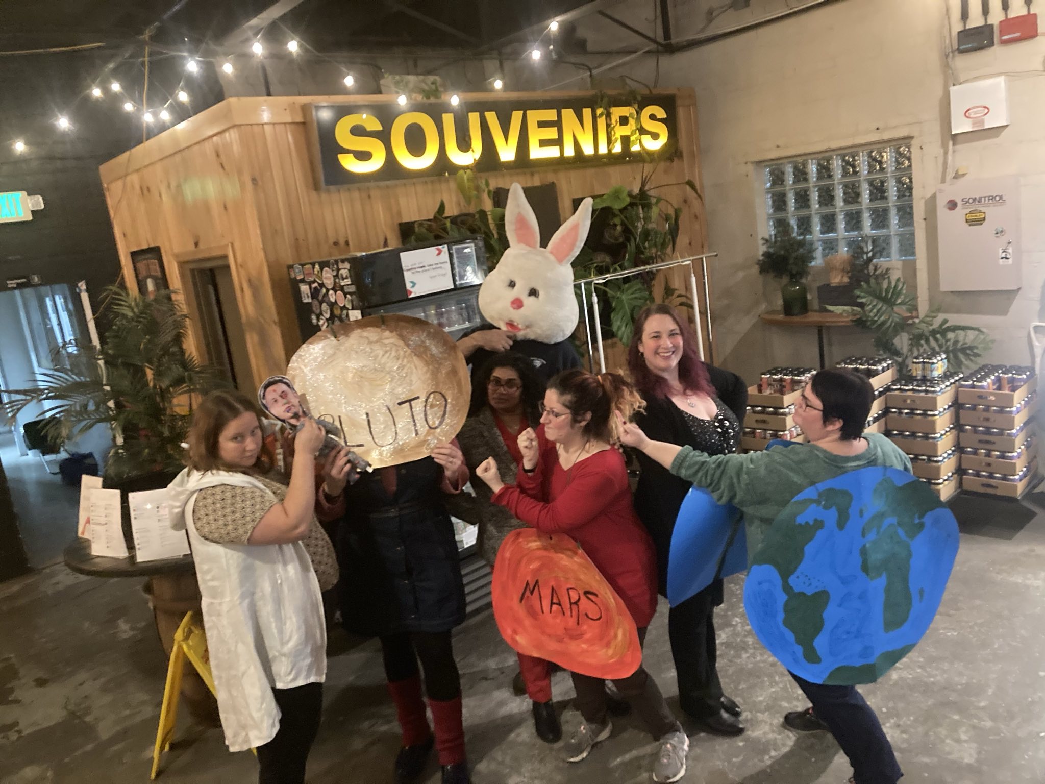 Group of children and adults in costumes, including a bunny and Earth, inside a souvenir shop with a sign reading 'SOUVENIRS'.