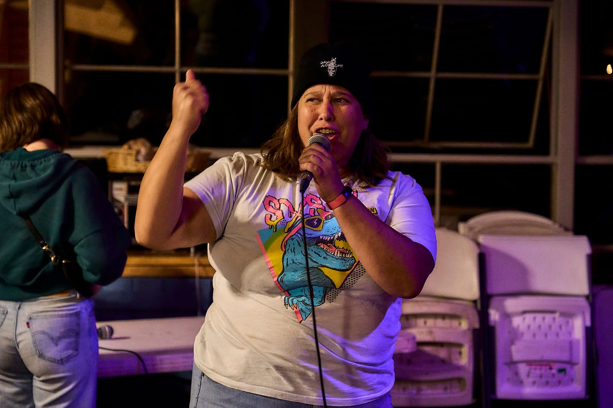 Person speaking into a microphone, wearing a beanie and a colorful T-shirt, gesturing with one hand, indoors at night.