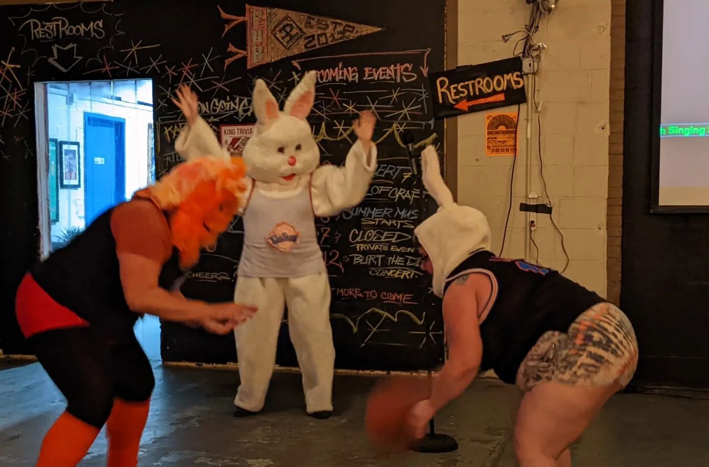 Two people with animal costumes and a person in casual clothing interact in a room with a blackboard and restrooms sign.