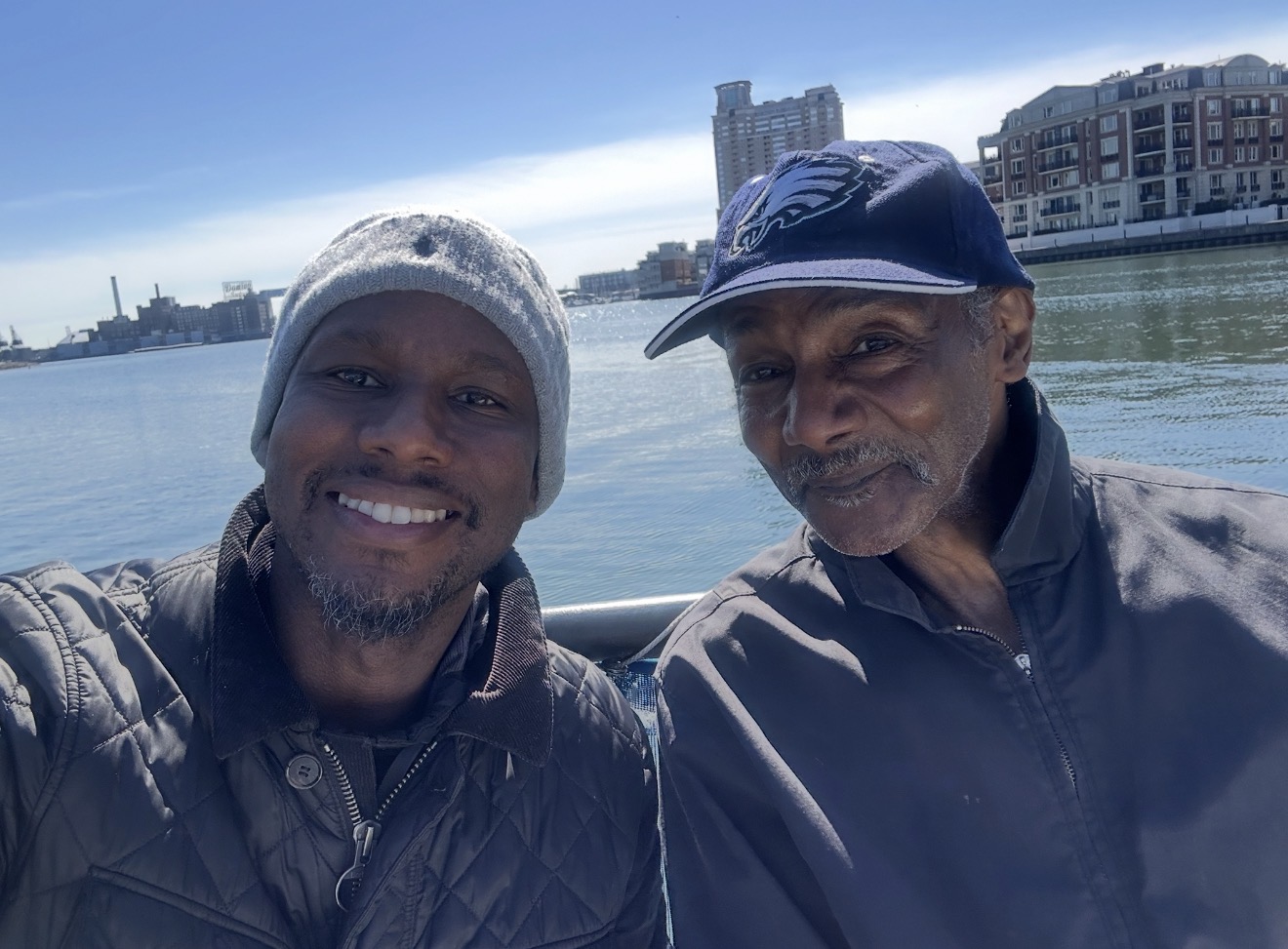 Two men smiling outdoors near water with buildings in the background.