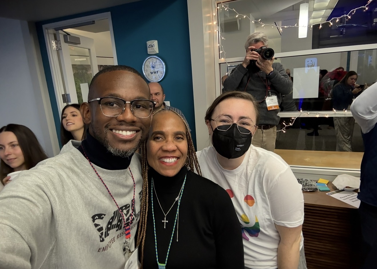 Three people smiling in front of a glass window, with a person taking a photo in the background.