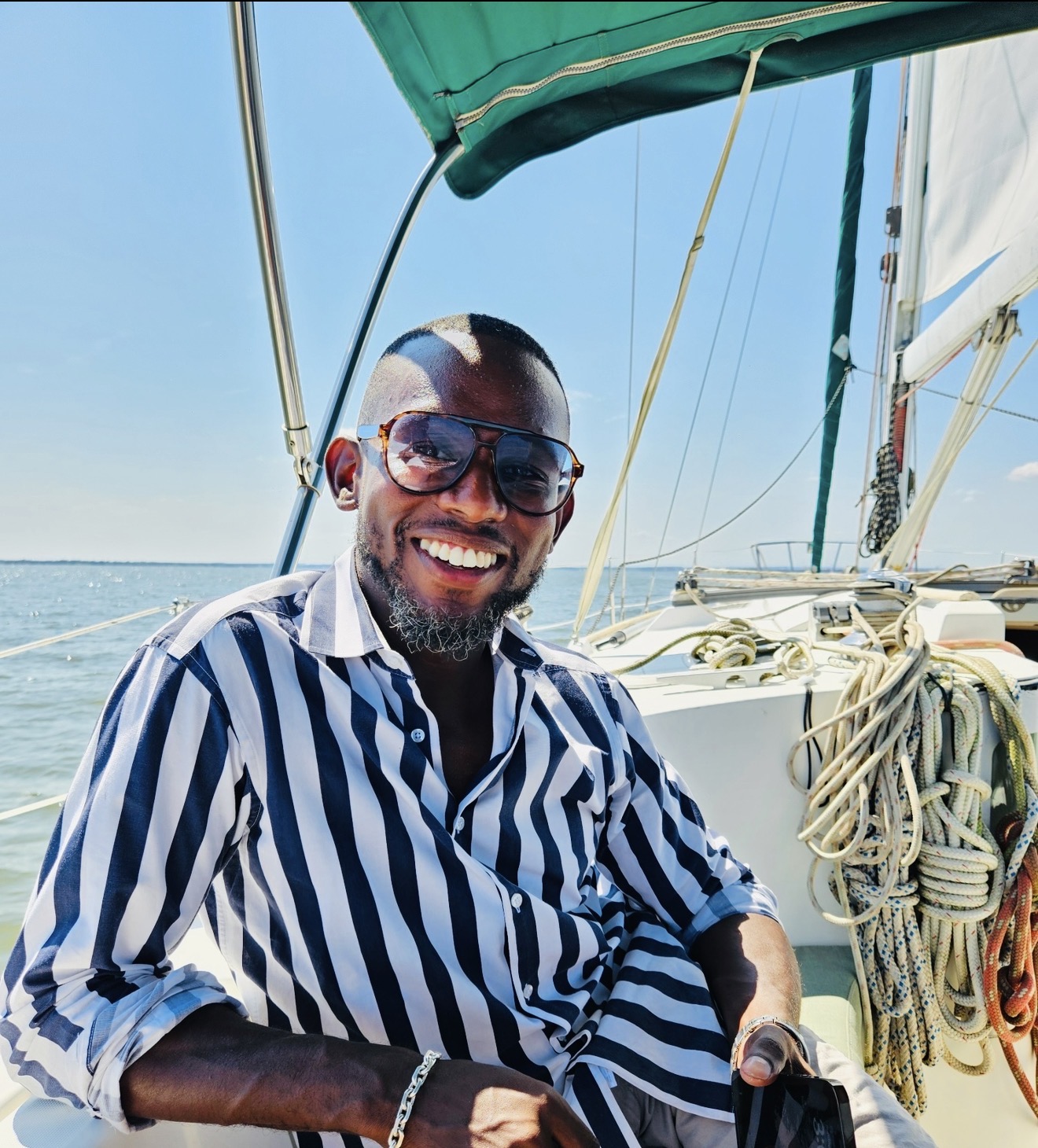 Smiling man with sunglasses on a sailboat, wearing a striped shirt, with water and sailboat rigging in background.