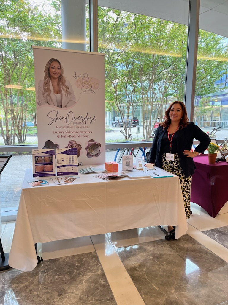 Woman standing next to a promotional table with a large poster inside a glass-walled building, trees visible outside.