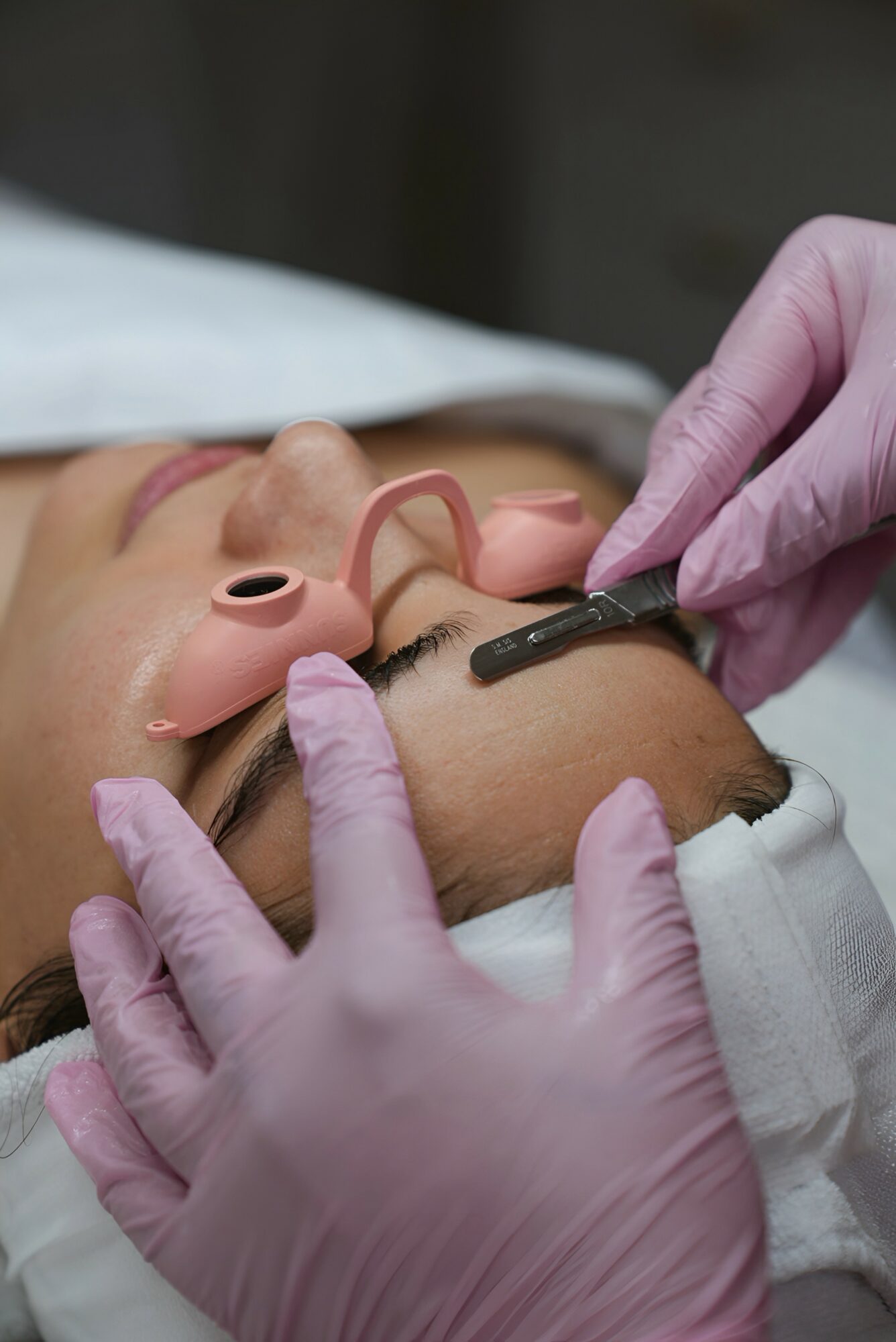 Medical professional in pink gloves performs a procedure on a patient's face with medical equipment, patient lying down with closed eyes.