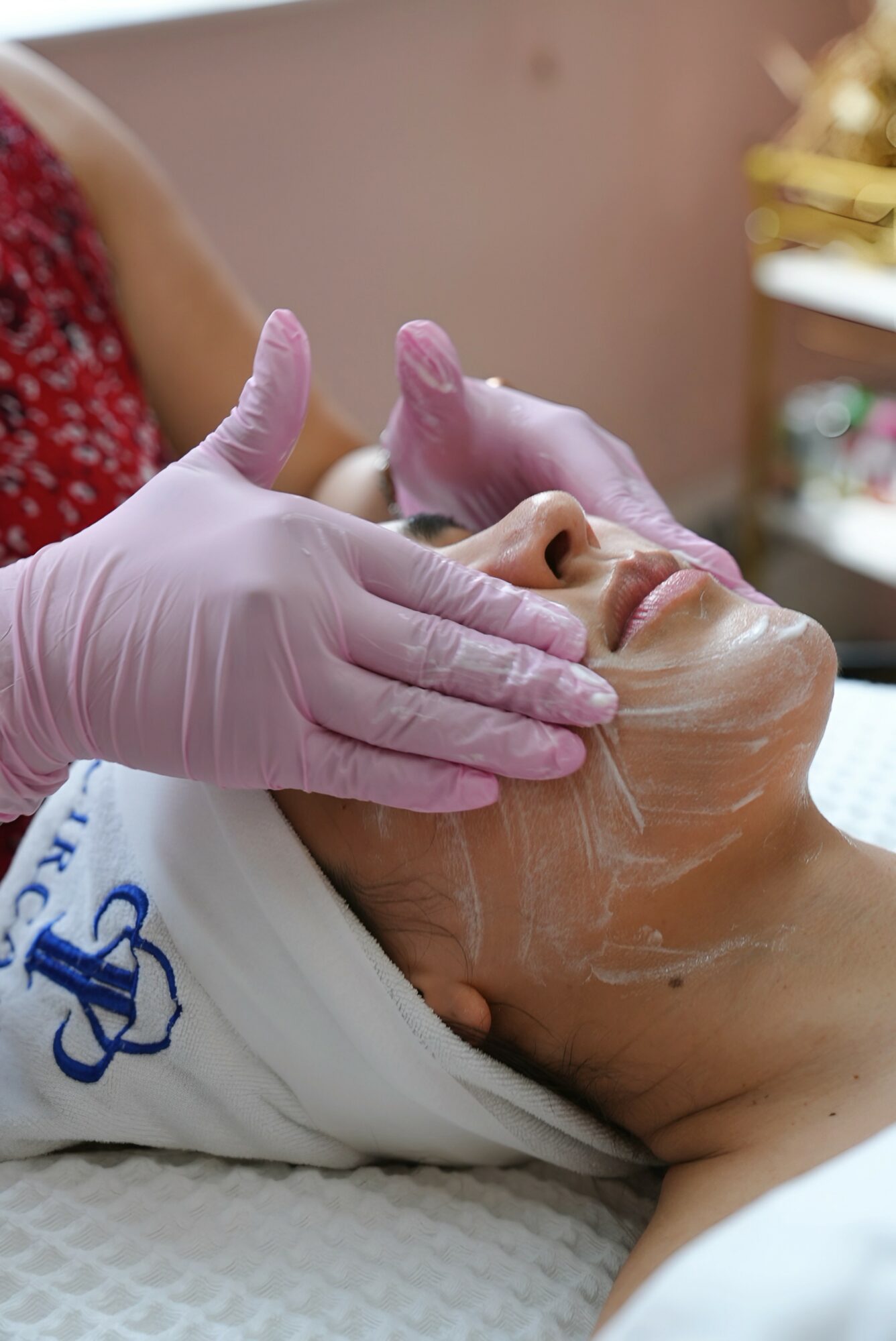 Person receiving facial treatment with gloved hands applying cream or mask, lying down with eyes closed.