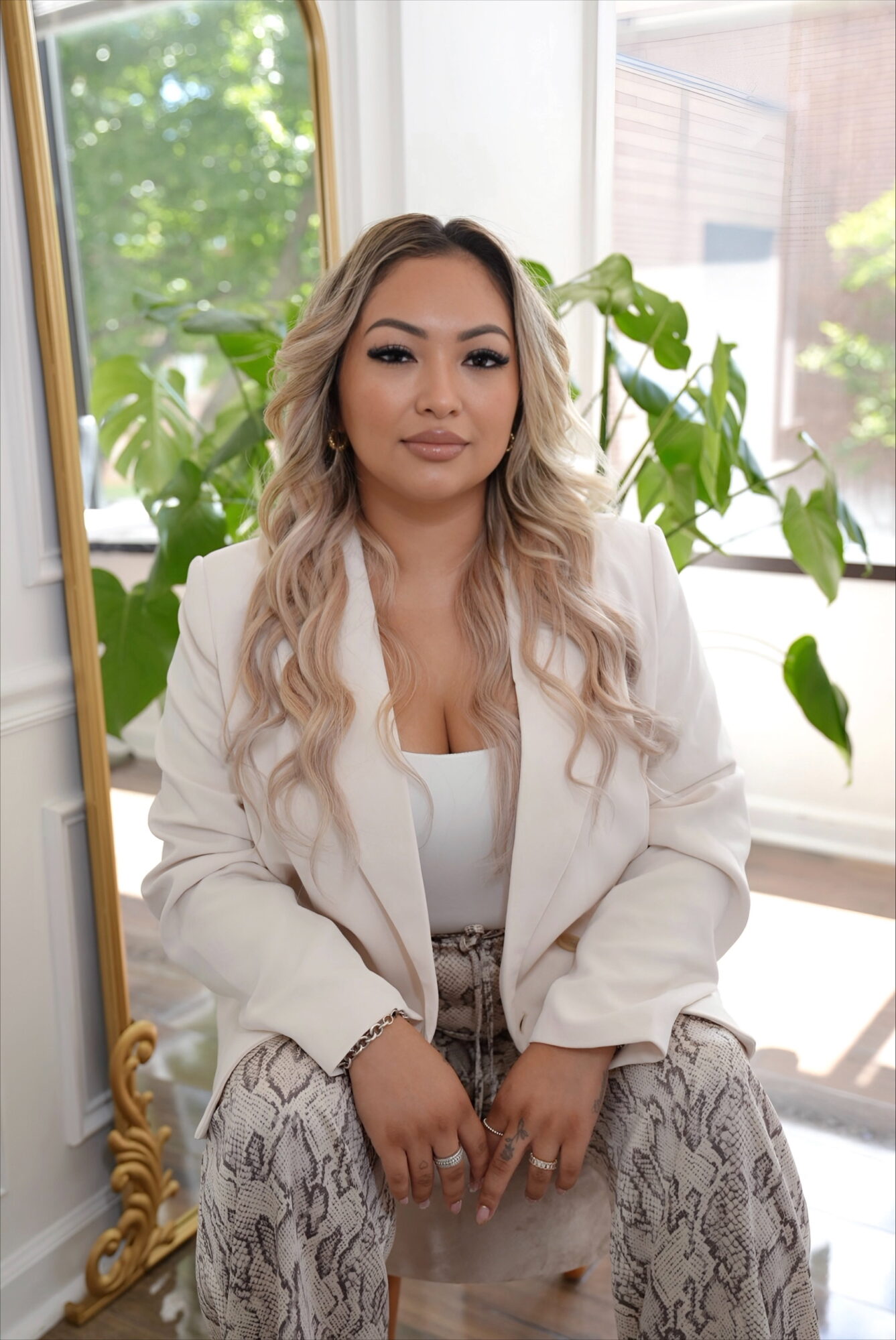 Woman with long wavy hair sitting indoors near a window with green plants, wearing a white blazer and patterned pants.