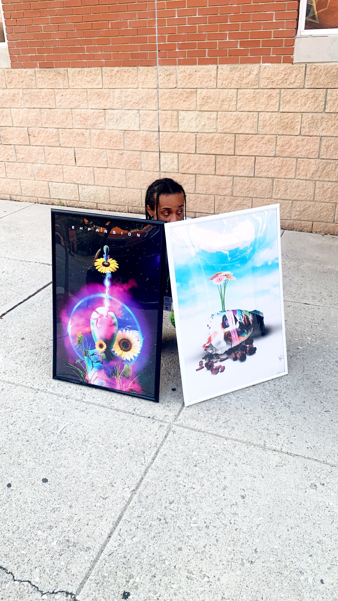Person holding two colorful paintings outdoors, one with a cosmic owl and the other with a pink flamingo on a boat, against a brick wall.