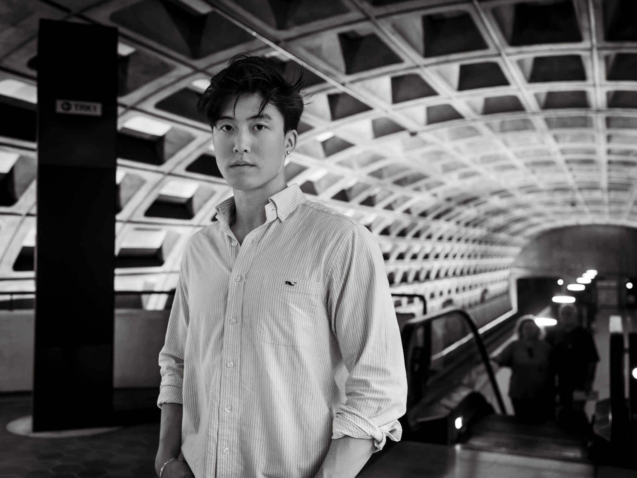 Young man standing in a modern underground station with geometric ceiling design, looking at the camera, in black and white.