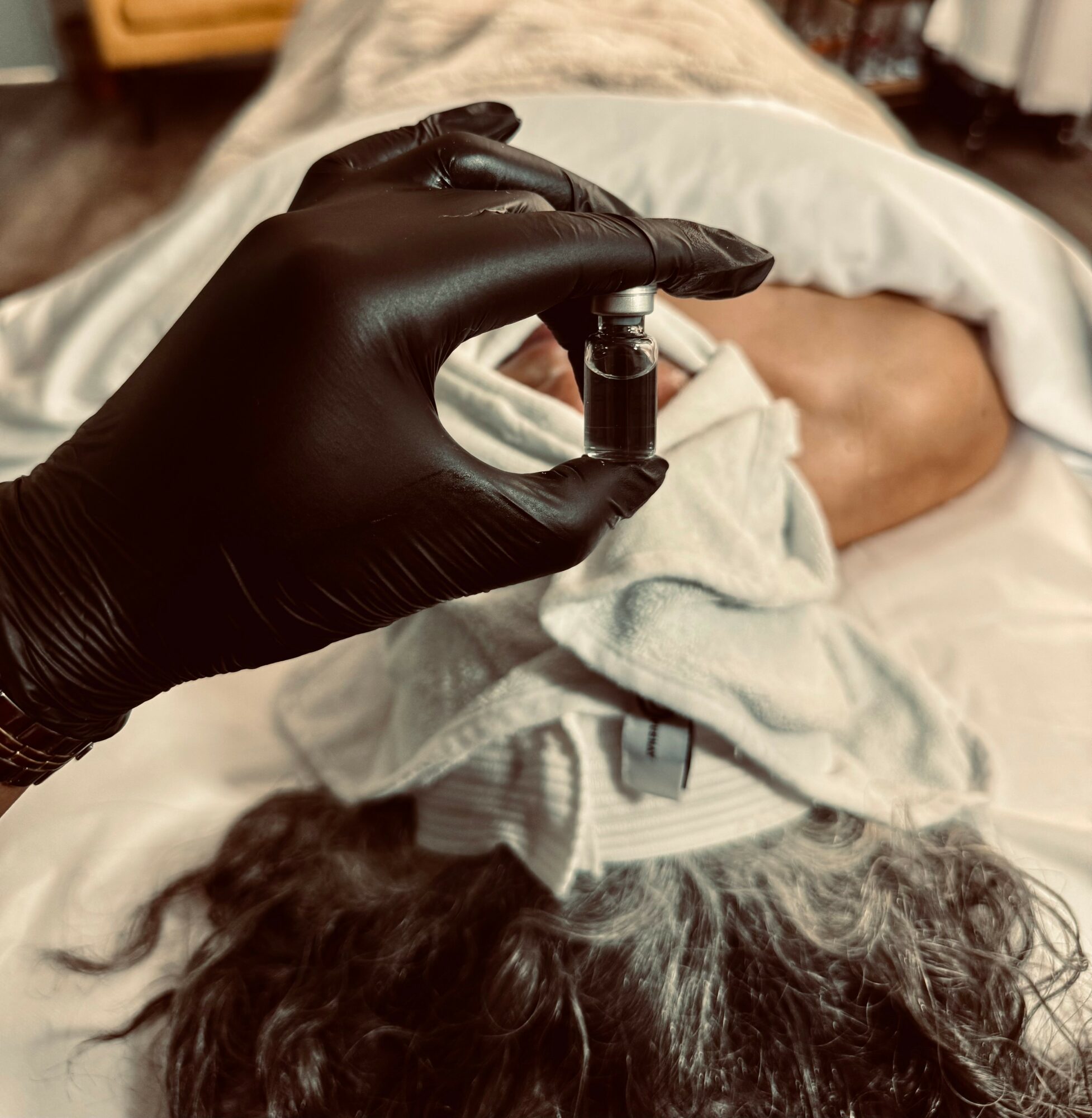 Person lying in bed holding a small vial or container, with a hand wearing a black glove, in a medical setting.