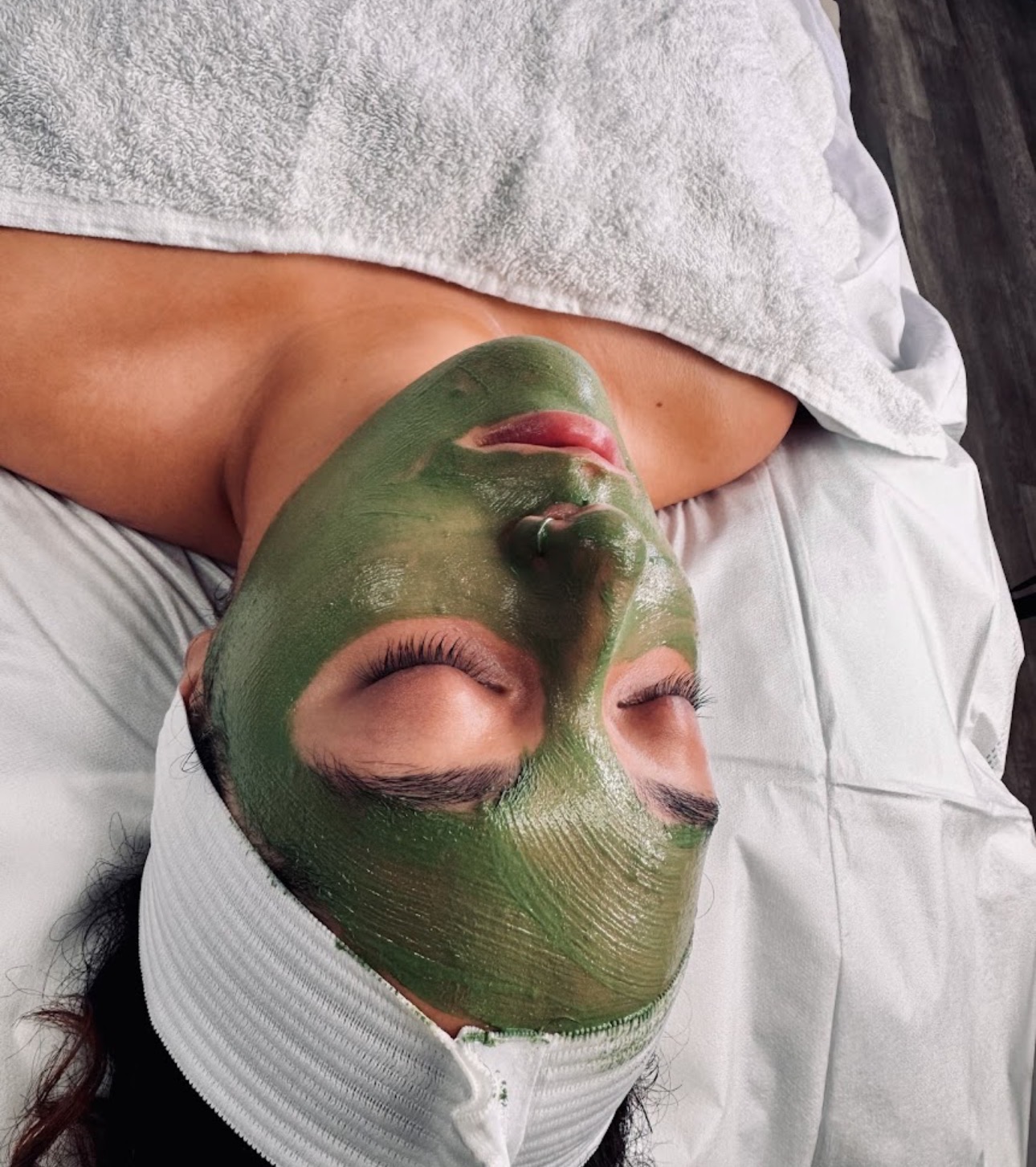 Person lying down with a green facial mask, eyes closed, wearing a white headband, on a bed with white sheets.