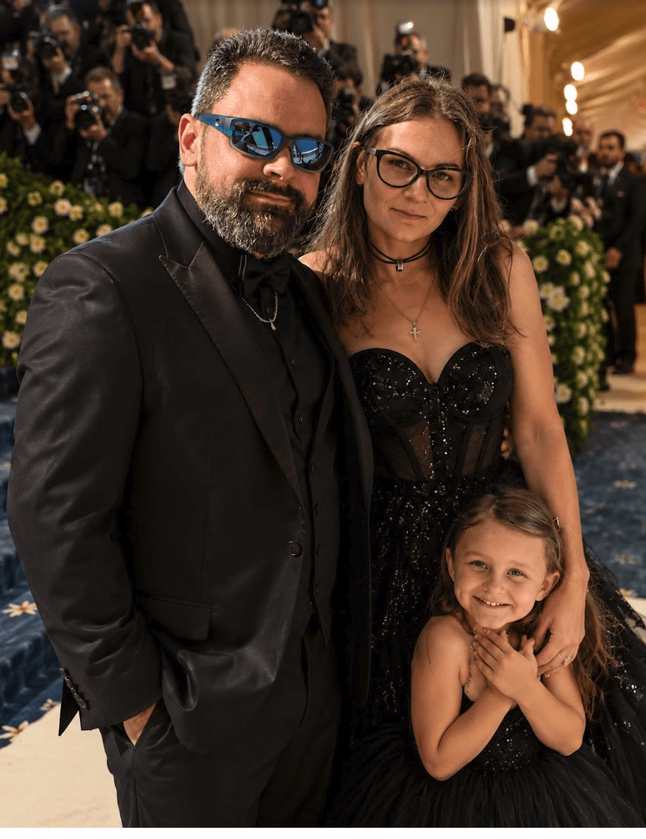 Three people, two adults and a child, pose together at an event, with a decorated background and crowd. 20 words.