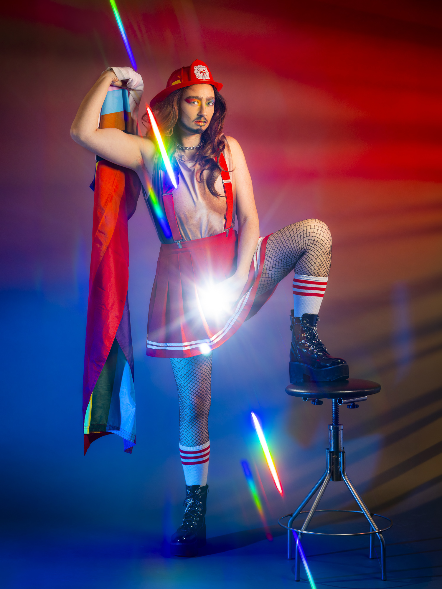 Person dressed in colorful costume with rainbow cape, fishnet stockings, and boots, standing on a stool with a rainbow light effect.