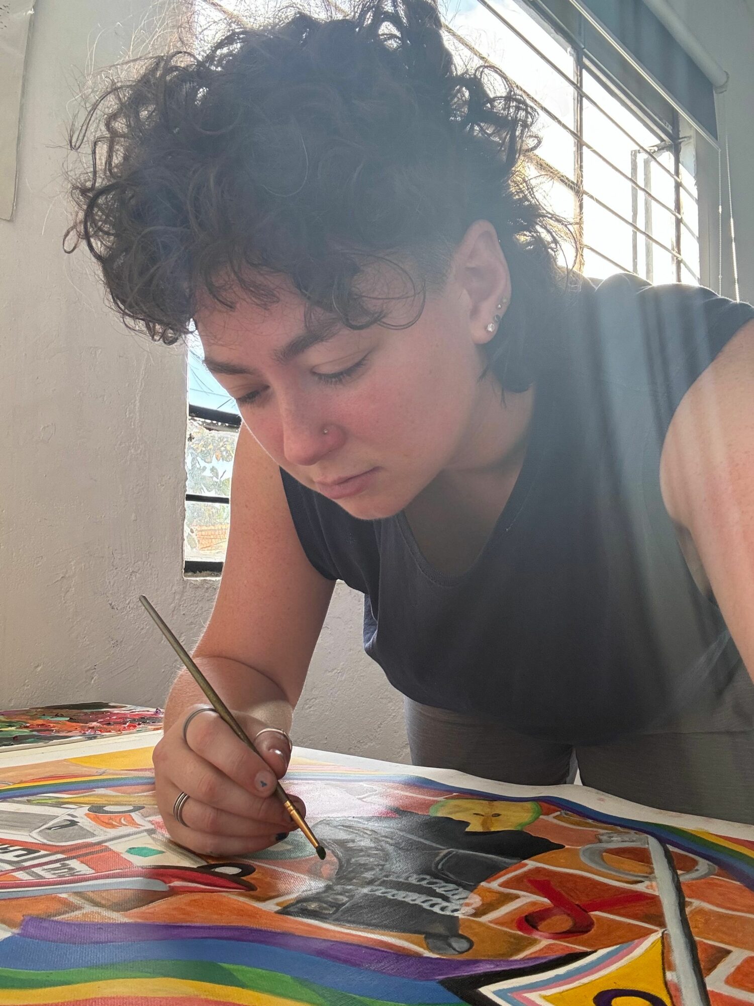 Person with curly hair painting on a colorful canvas, sitting indoors near a window.