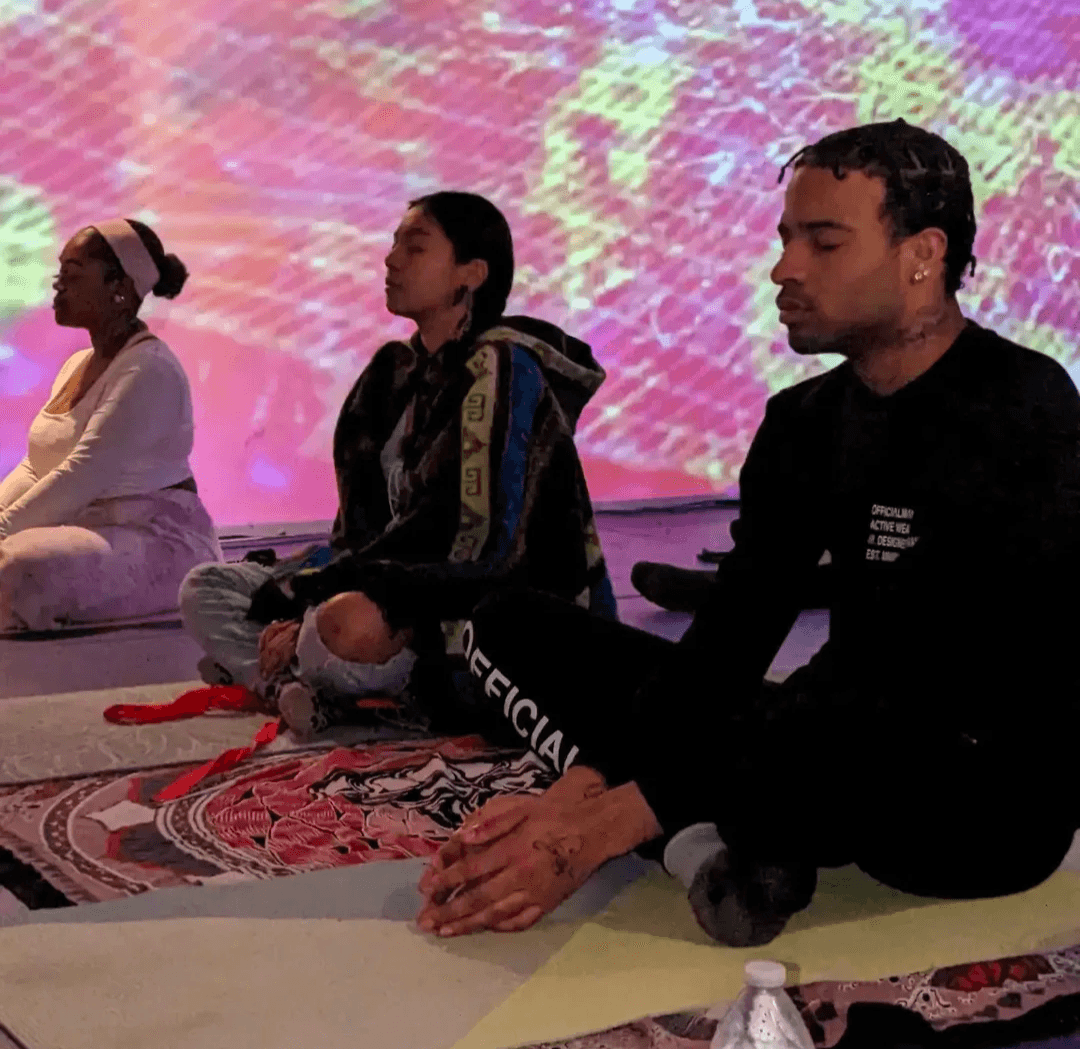 Three people sit cross-legged on a patterned rug with a colorful background, meditating or practicing mindfulness.