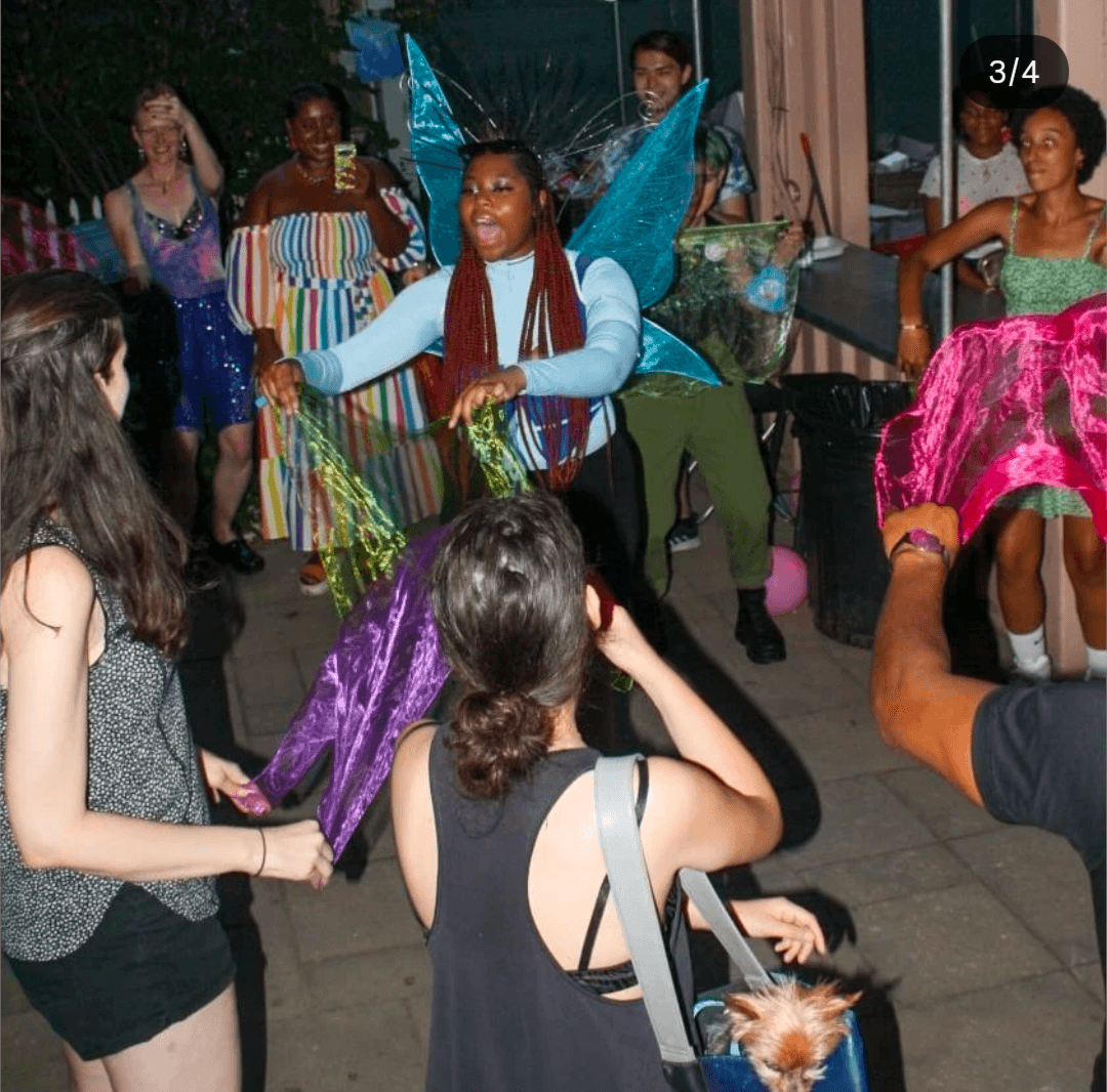 Group of people dancing outdoors, some in colorful costumes, with a woman in the center smiling and holding hands.