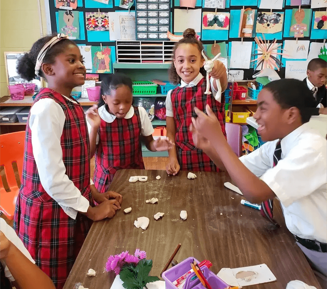 Four children in school uniforms are playing with clay around a table in a classroom, smiling and laughing.