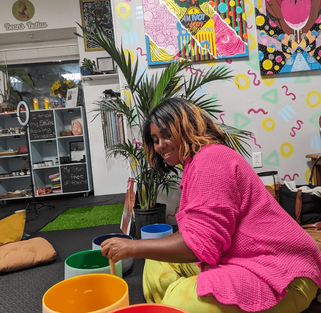 Woman with curly hair in a pink sweater sitting on the floor with colorful bowls, plants, and artwork behind her.