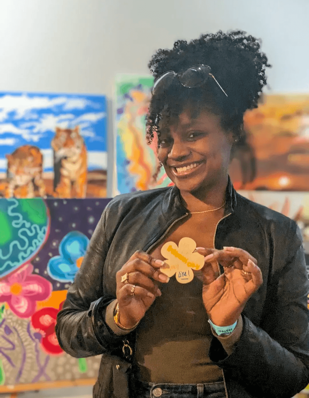 Woman with curly hair smiling, holding a small object, standing in front of colorful paintings.