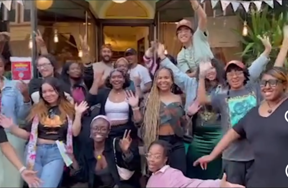 Group of people smiling and waving outside a building, some raising hands, in a lively setting.