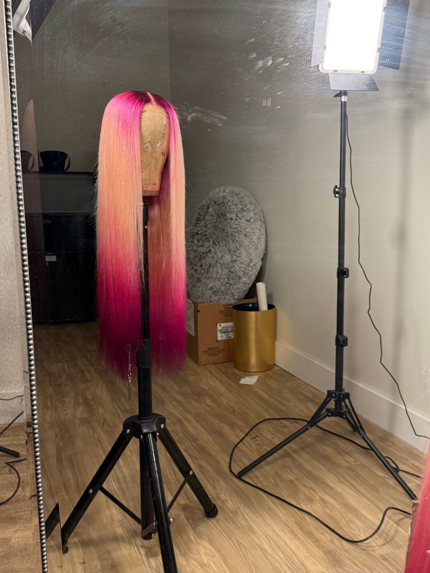 Pink and blonde hair extension on a stand, with a gray foam head, gold container, and lighting equipment in the background.