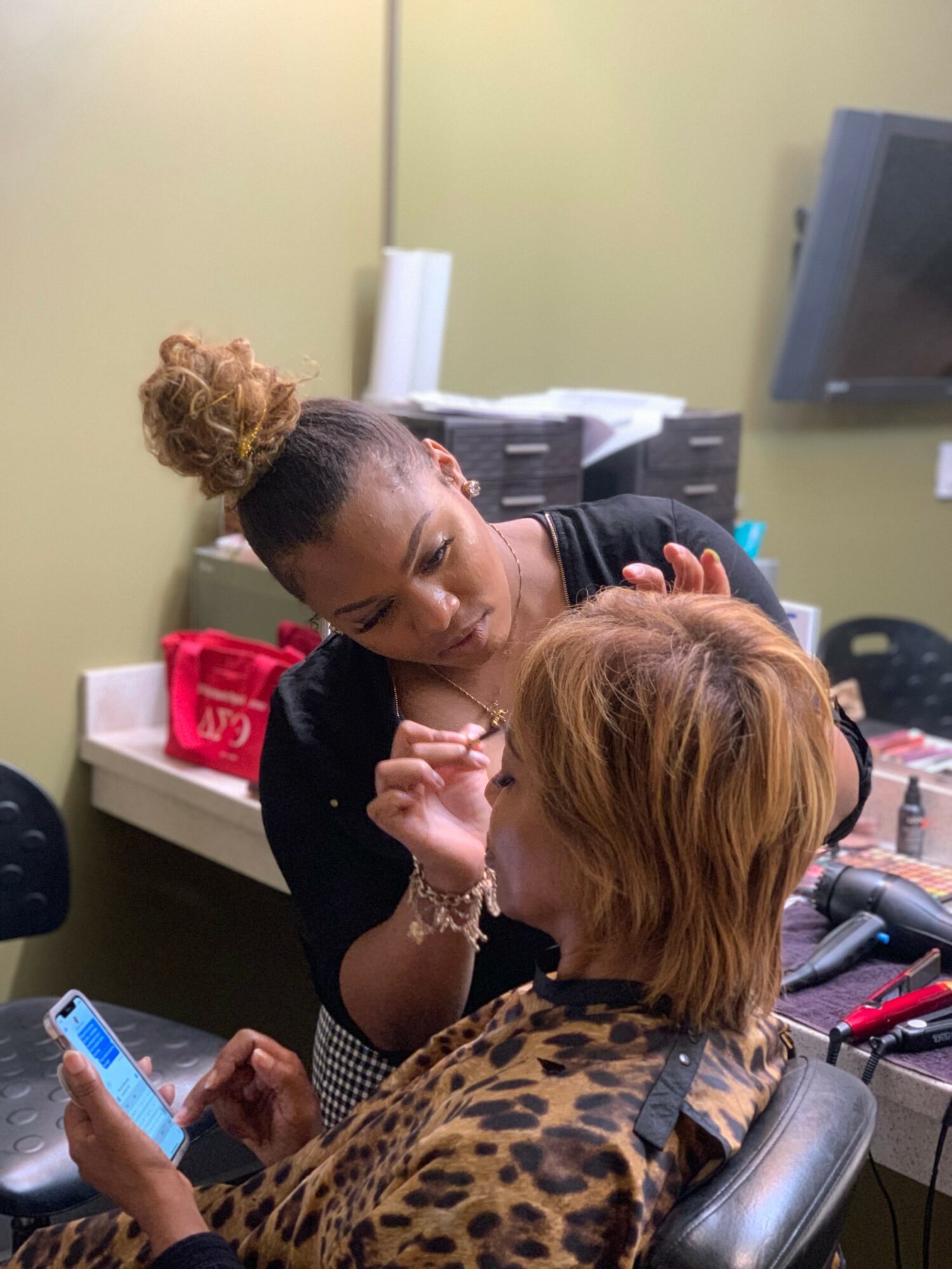 Makeup artist applies makeup to a person with short, reddish hair in a room with a green wall and a television.