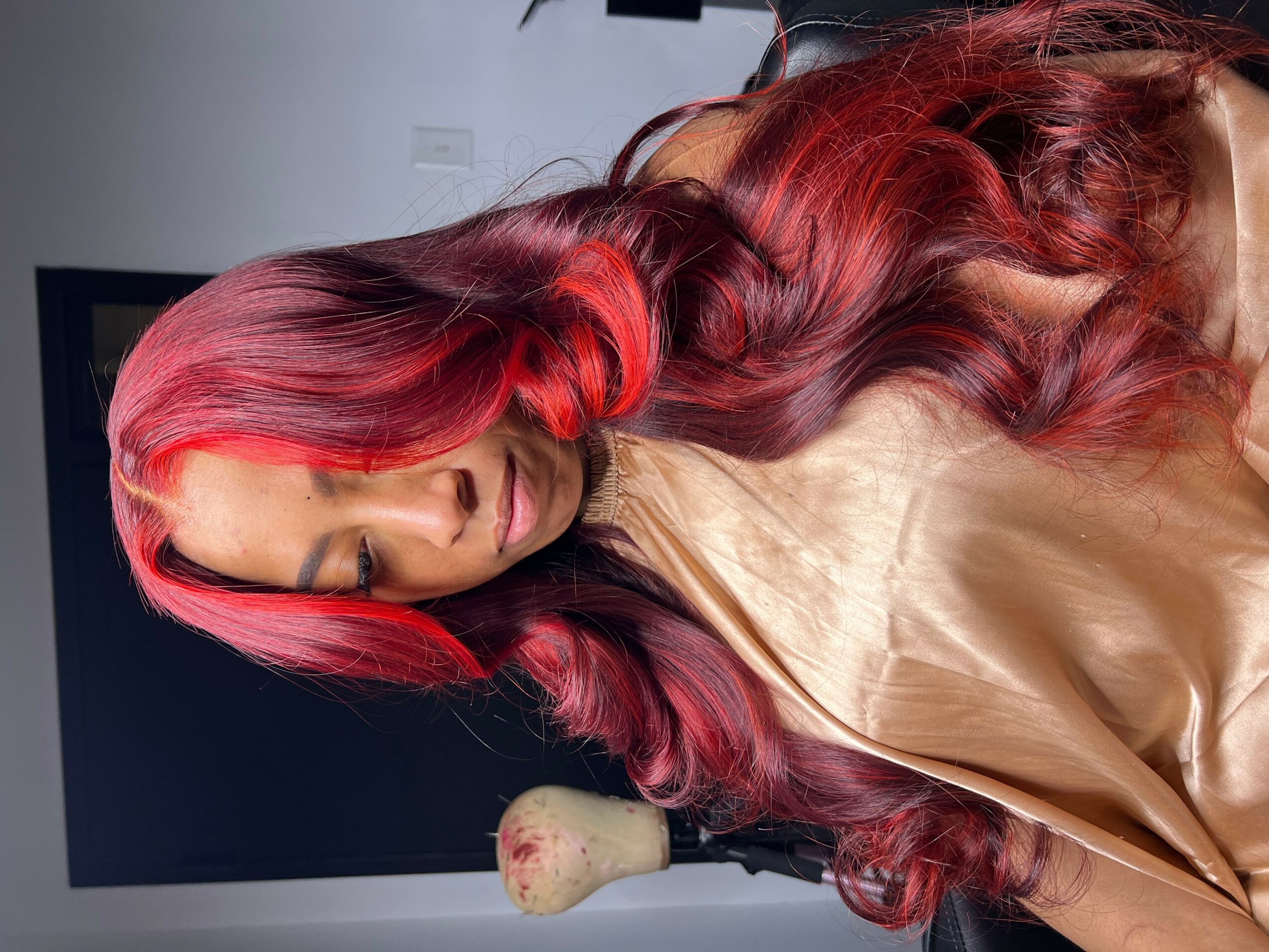 Woman with long, wavy red hair looking down, wearing a beige top, in a room with a dark background.
