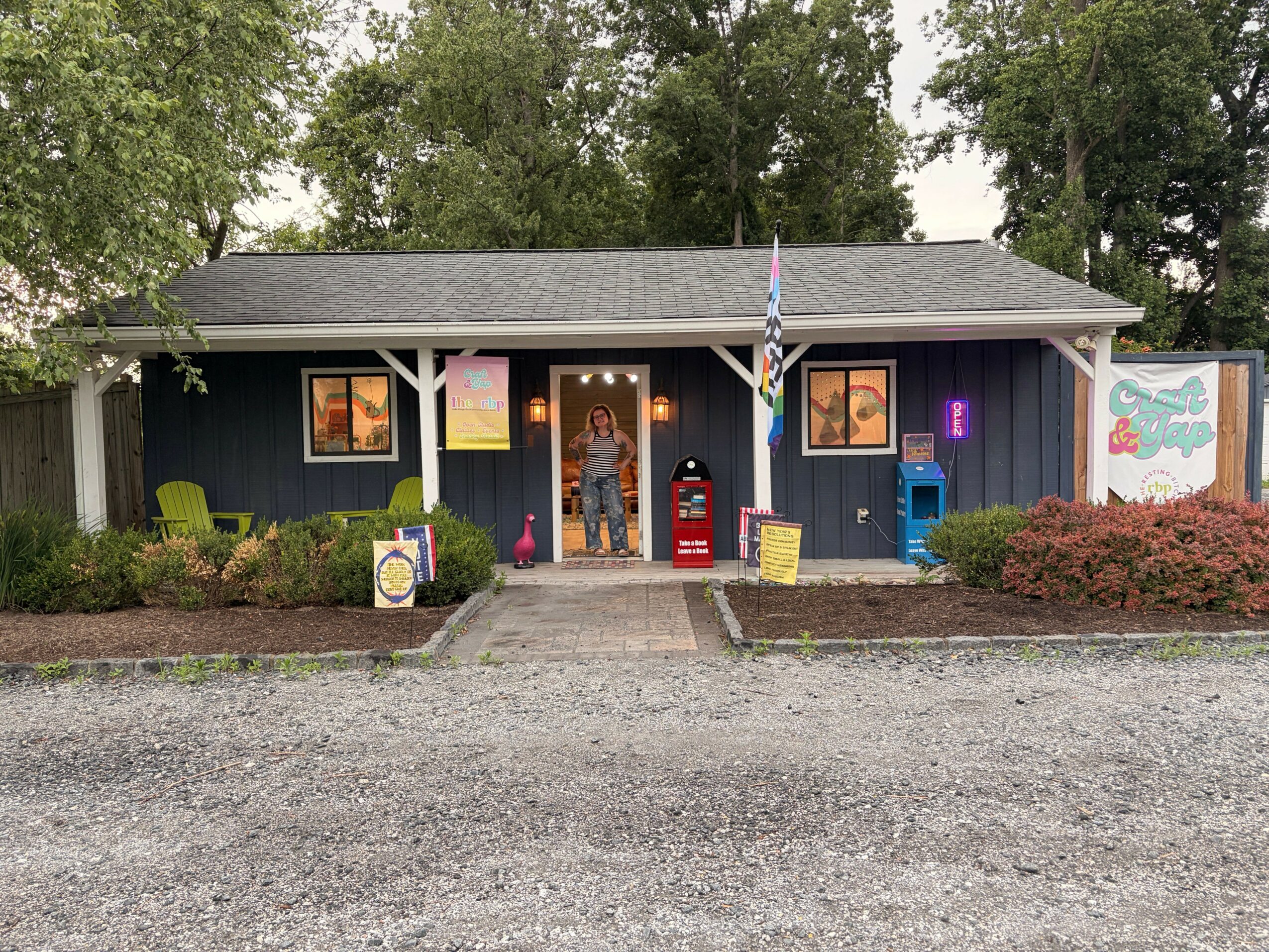 Gallery with paintings, a red car, and a flag outside, viewed from the side, with trees and a gravel path.