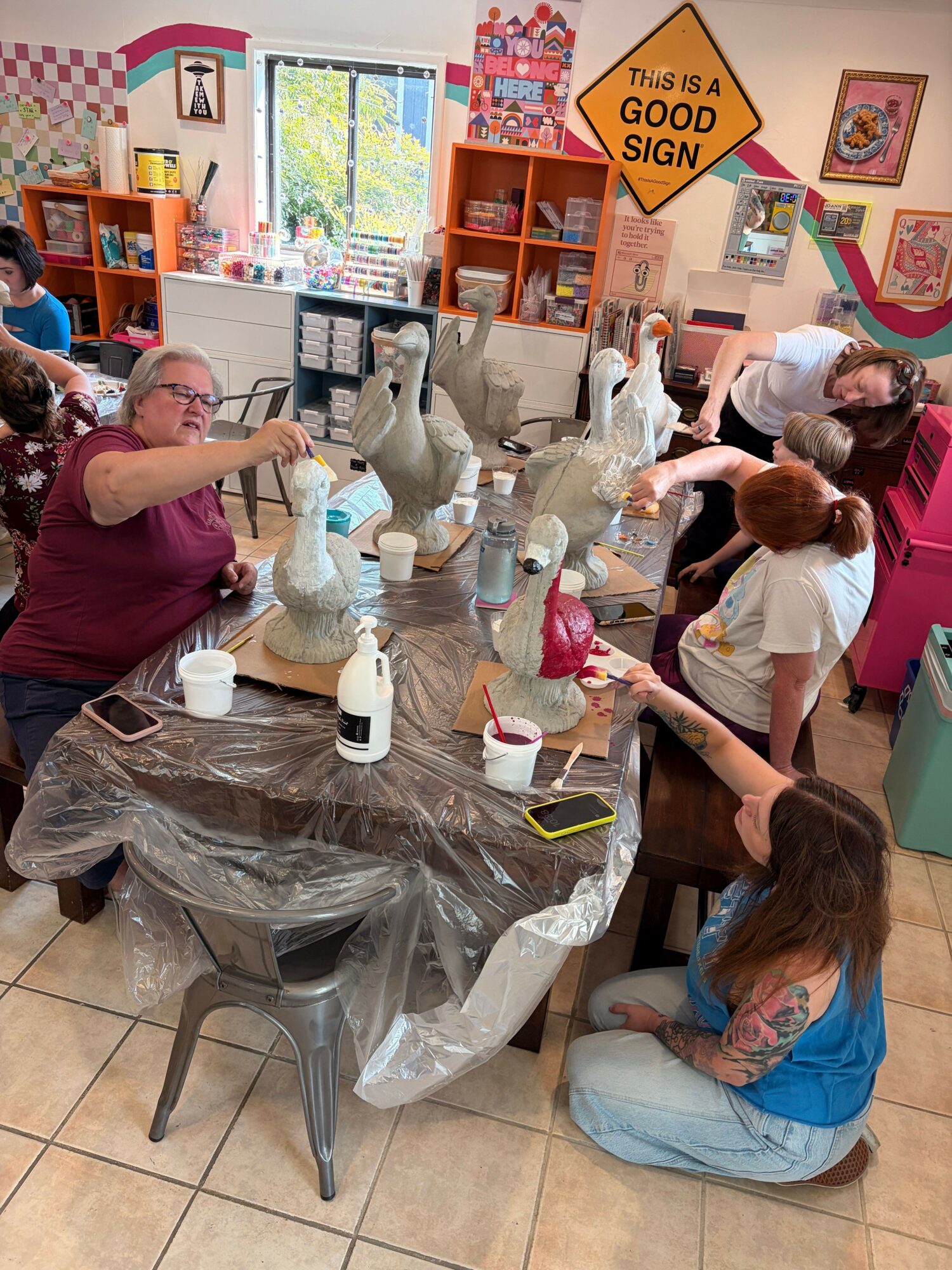 Group of people working on art projects at a table in a colorful room, with art supplies and decorations visible.