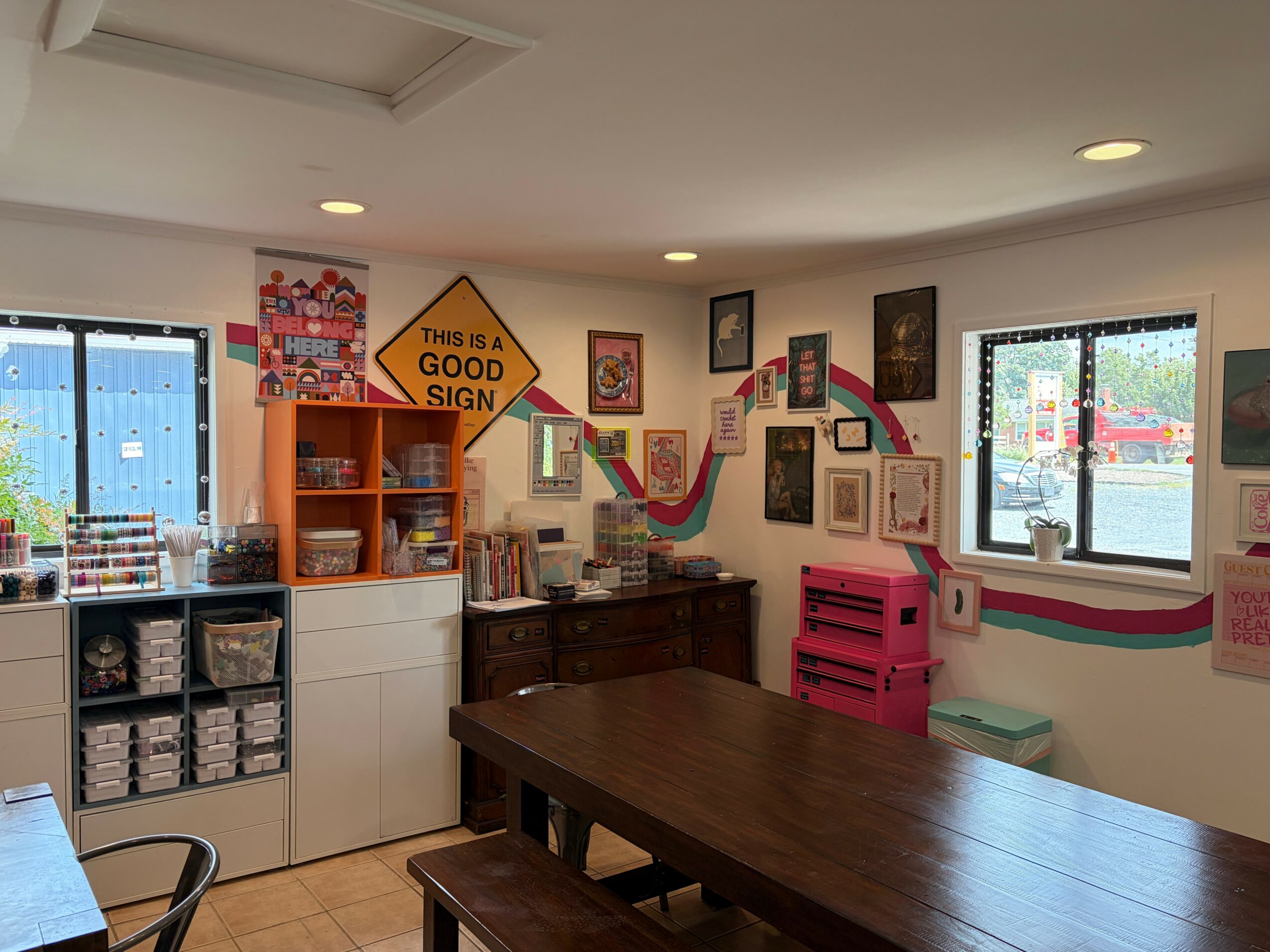 Room with colorful wall art, window, wooden table, and storage units, decorated with rainbow and framed pictures.