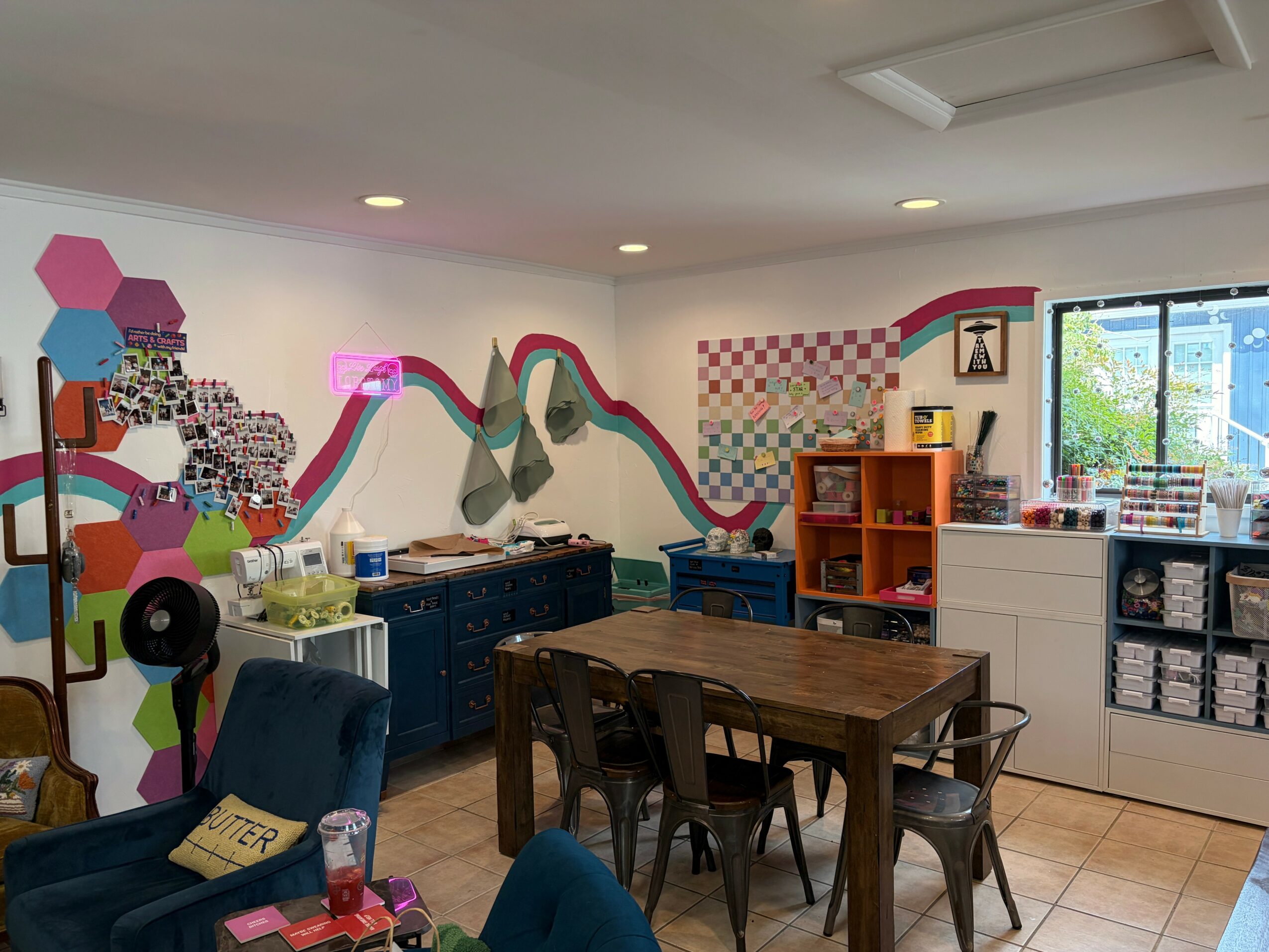 Room with colorful wall art, a wooden table, chairs, and storage shelves near a window.