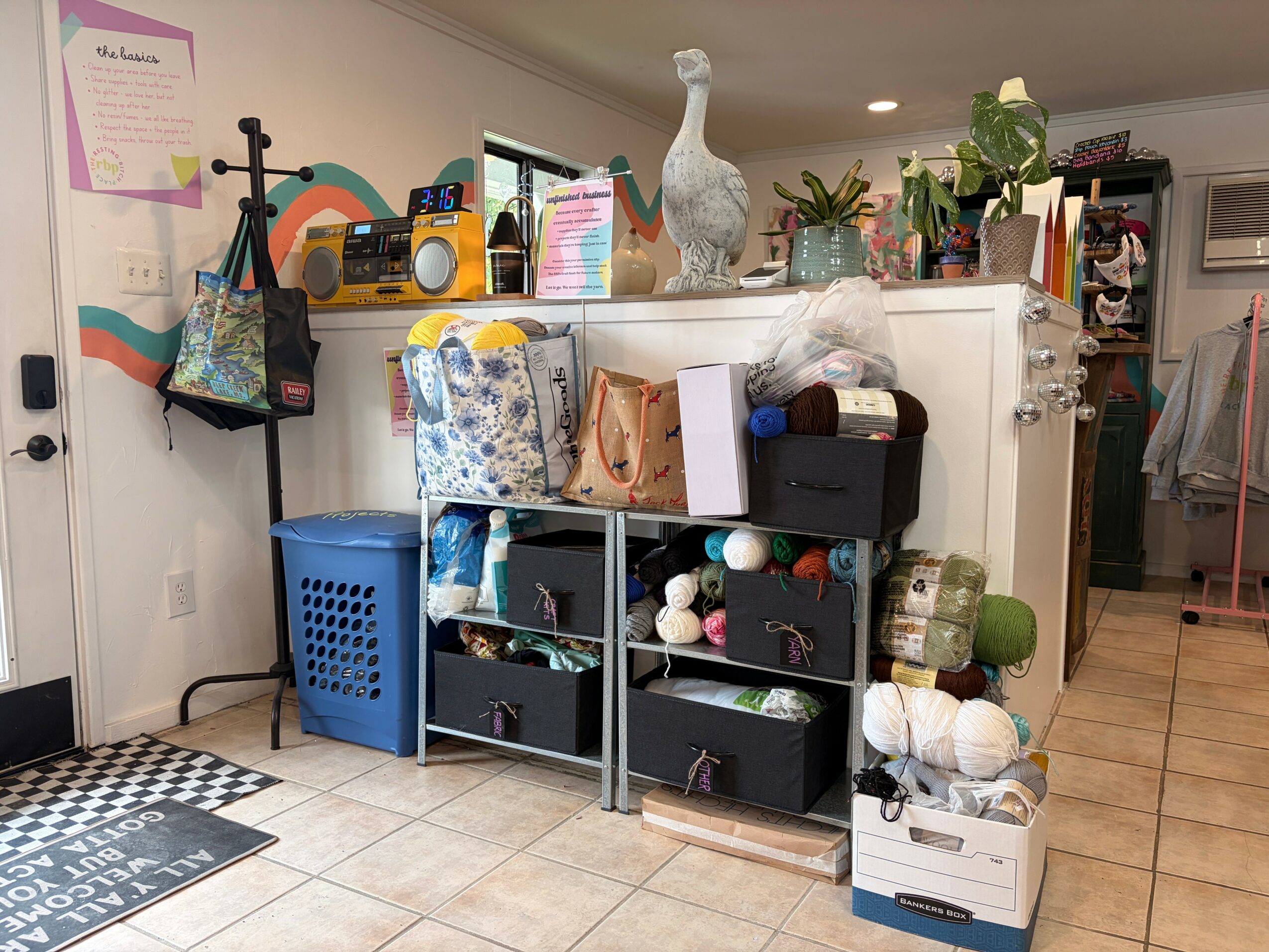 Cluttered room with shelves, plants, and various objects on a white counter, with a blue laundry basket nearby.