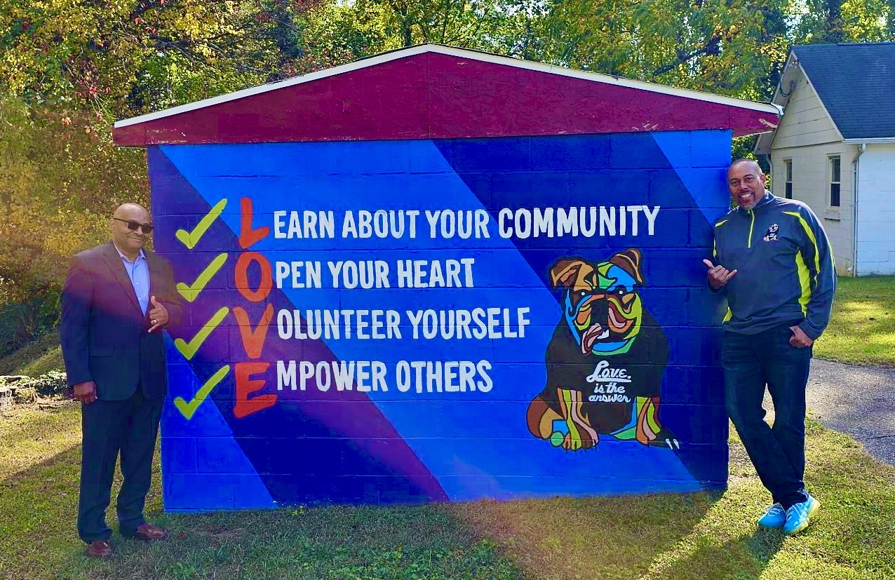Two men stand beside a colorful community banner with a tiger illustration, promoting community involvement and empowerment.