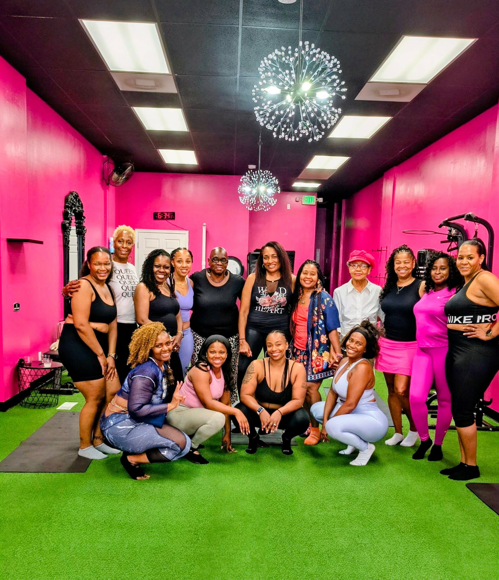 Group of women in fitness attire posing in a pink and black gym with green flooring, fitness equipment, and ceiling lights.