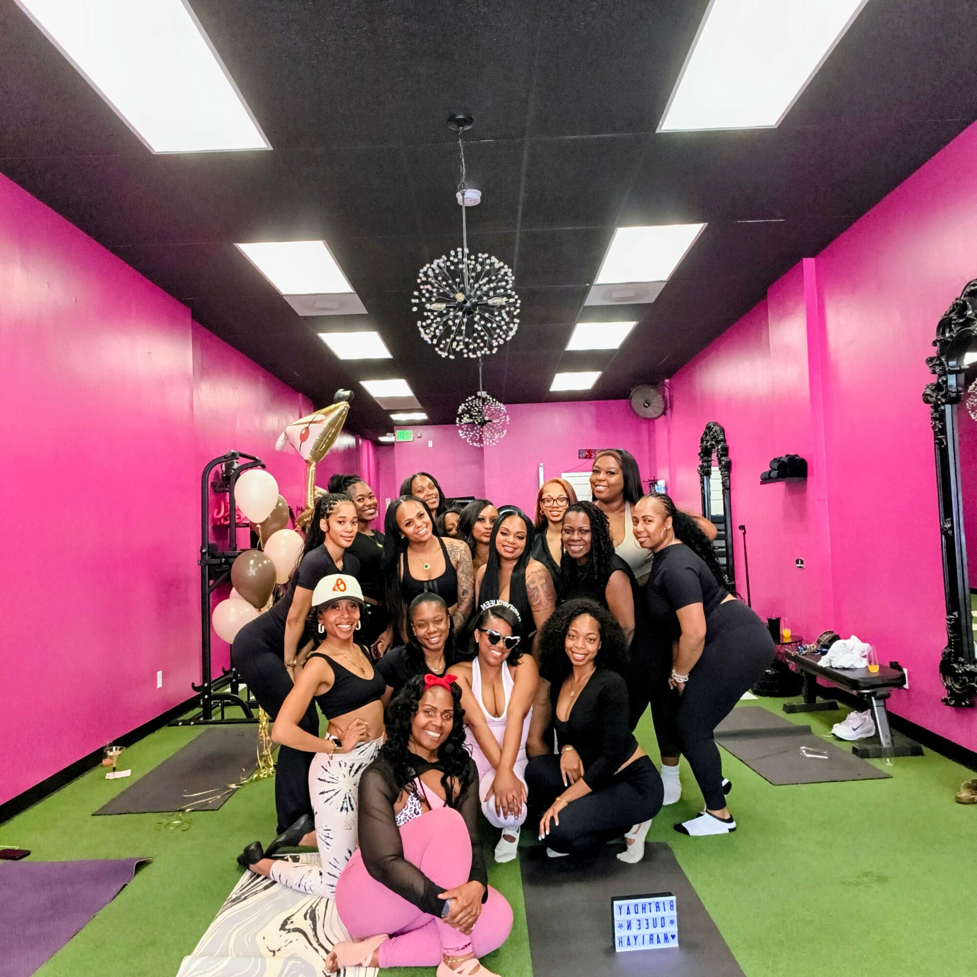 Group of women posing in a brightly colored room with pink walls, mirrors, and green flooring, some wearing black outfits and glasses.