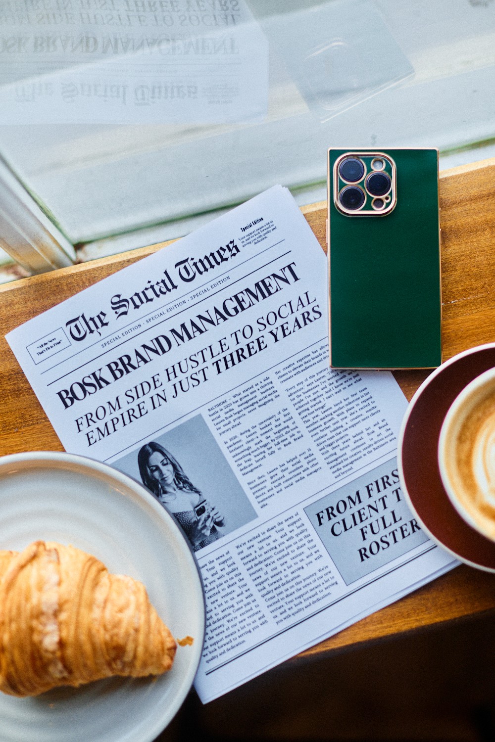 Newspaper on table with croissant, coffee, and smartphone near window, sunlight illuminating the scene.