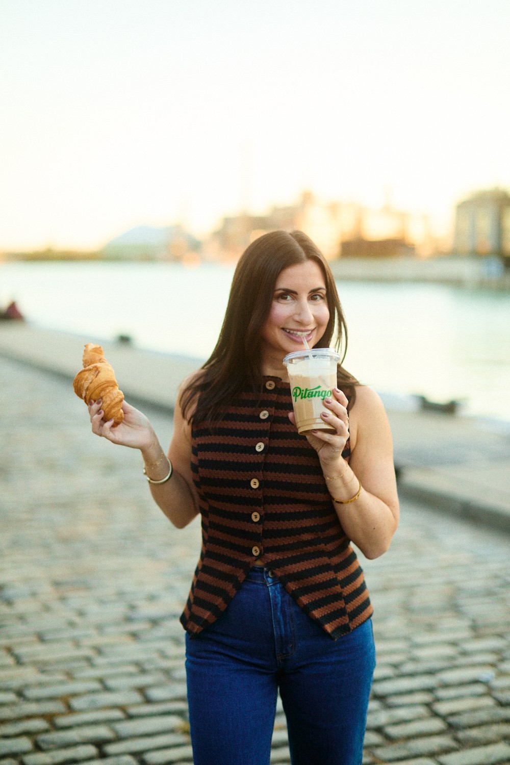Woman holding a croissant and a drink outdoors near water, smiling, with buildings in the background.