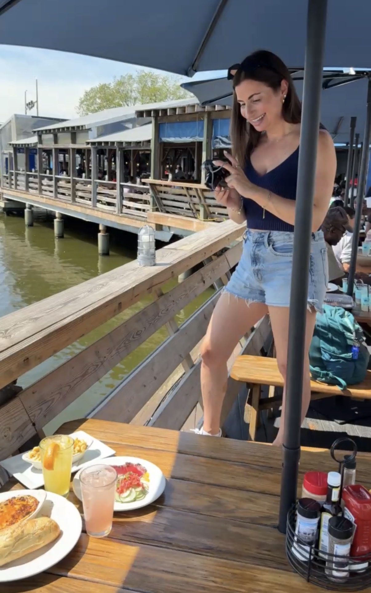 Woman standing by a railing, looking at her phone, with a waterfront restaurant in the background, and food on the table.