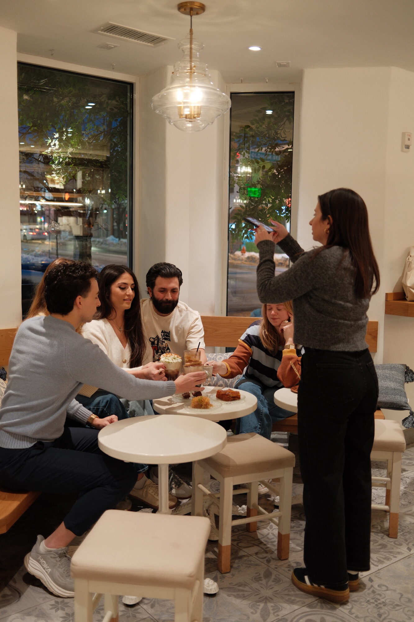 Group of five people sitting around a table in a restaurant, woman standing and talking, chandelier overhead, large windows showing street view.