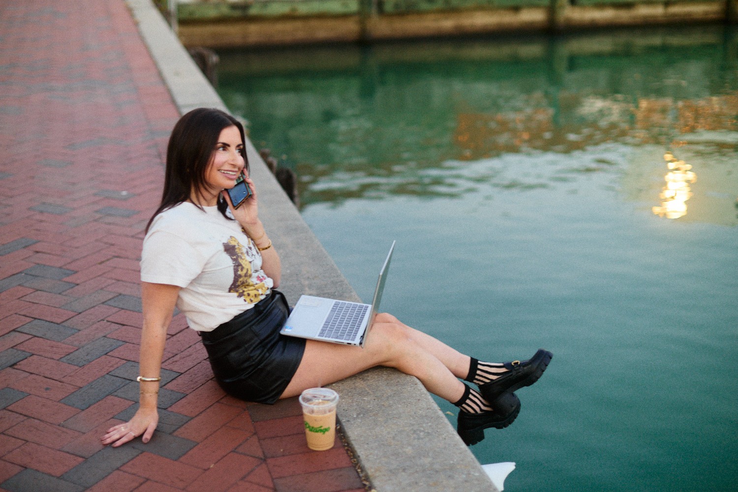 Woman sitting on brick walkway by water, using laptop and talking on phone, with a drink nearby.