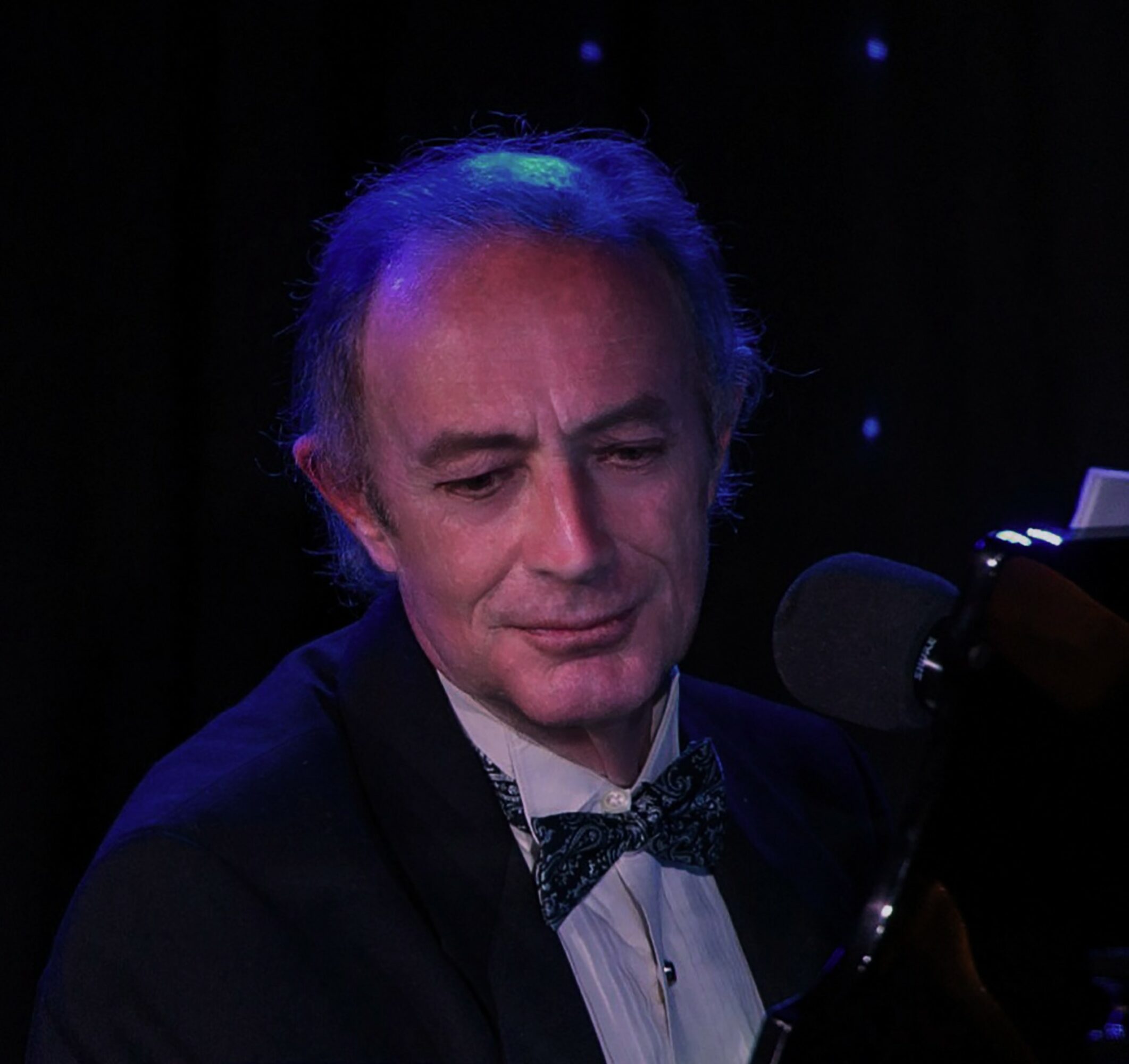 Man with gray hair in formal attire, wearing a bow tie, sitting at a piano with a microphone, dark background.