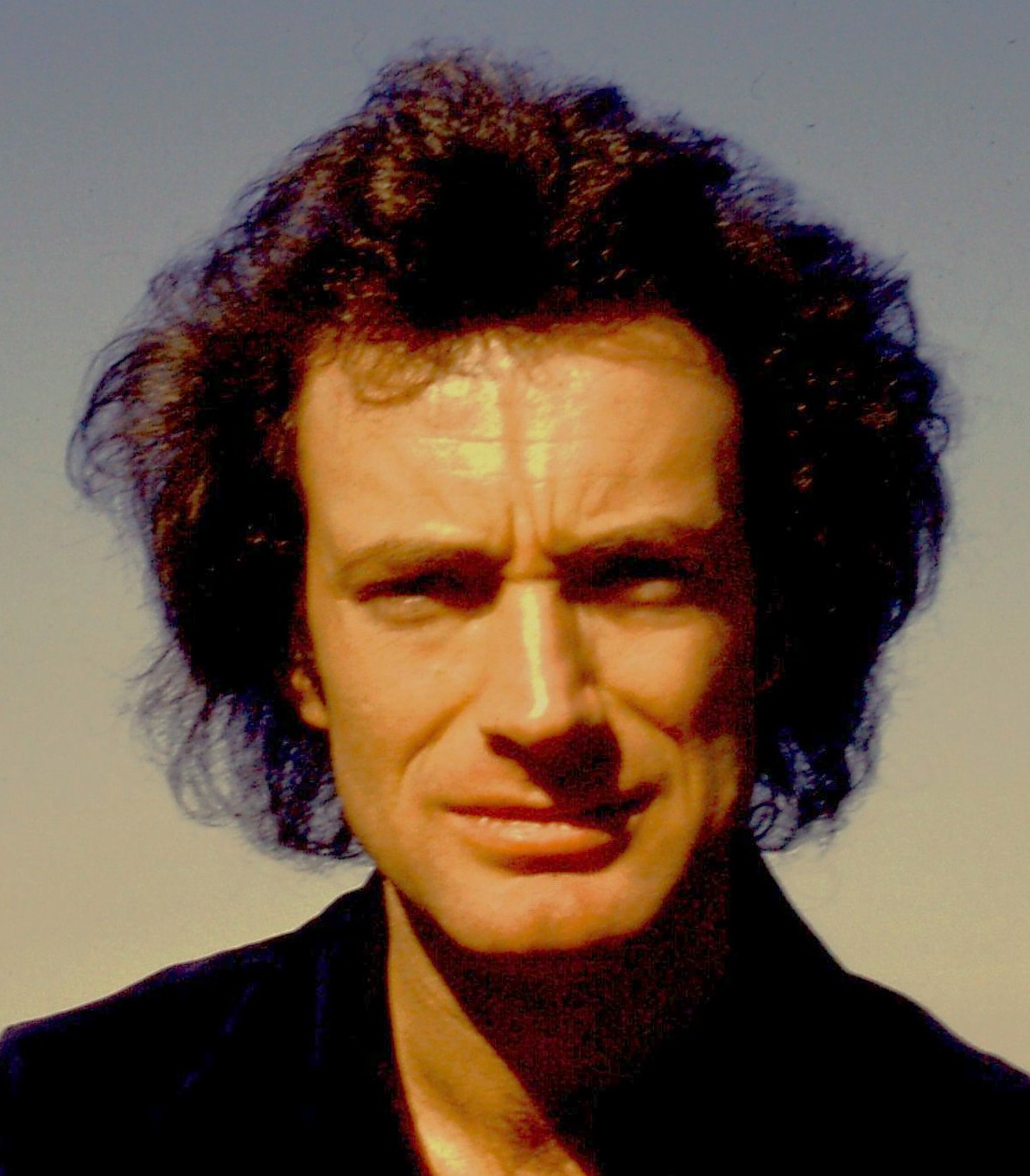 Portrait of a man with curly hair and a serious expression, against a clear sky background.