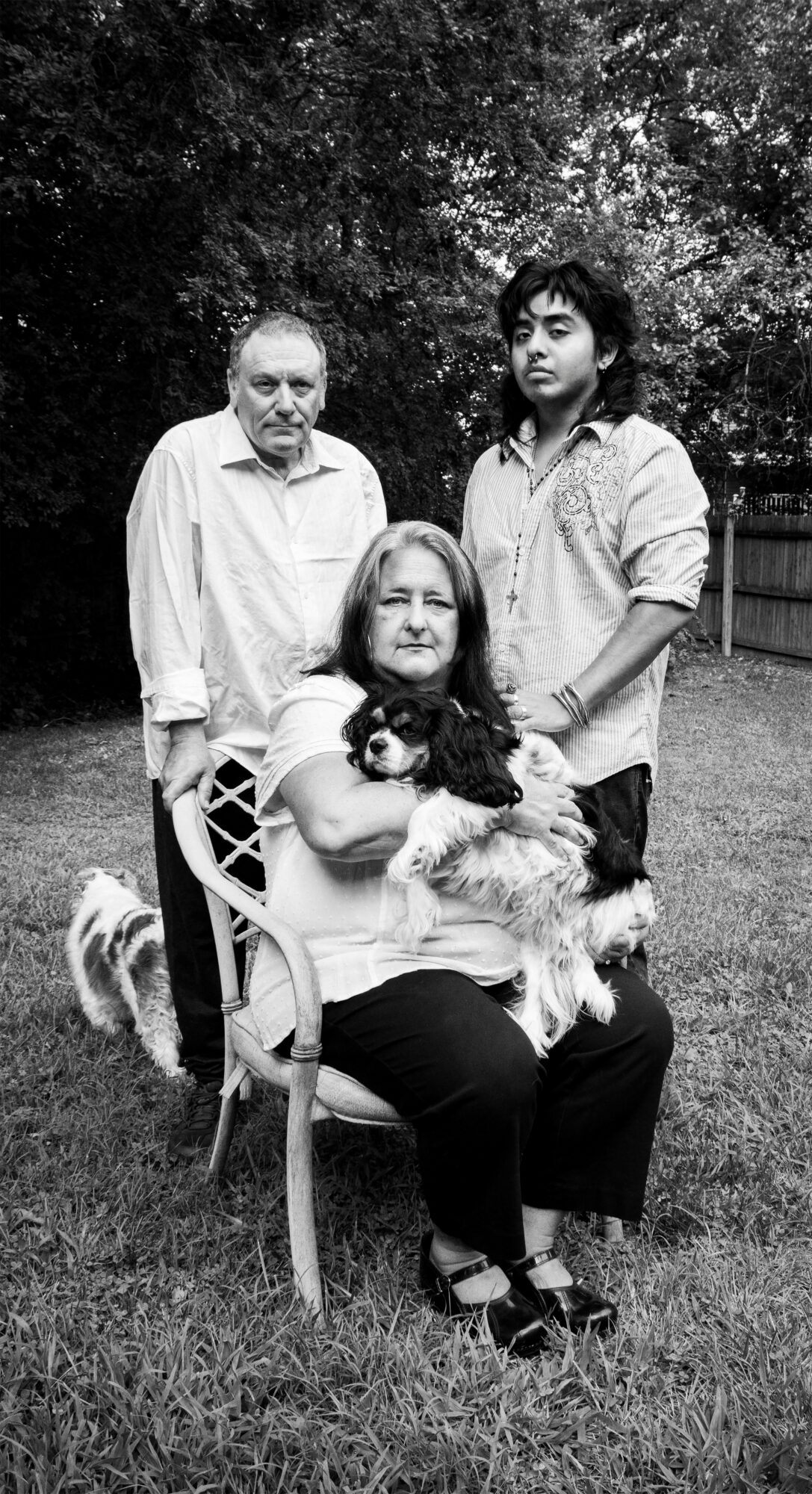 Three people outdoors, one sitting on a chair holding a dog, two standing behind, trees in background.