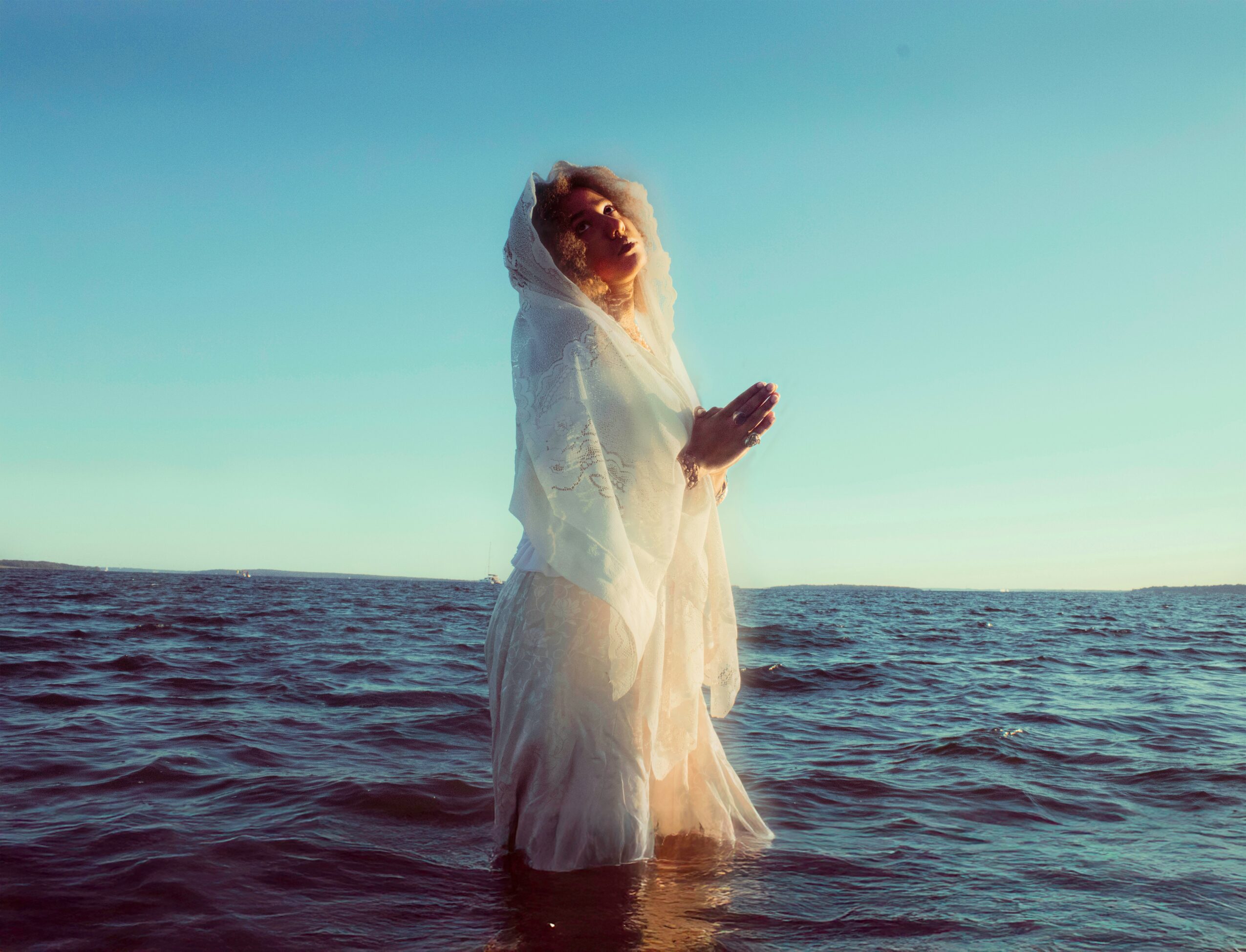 Person standing in water at the ocean, wearing a white dress and veil, with a clear sky background.