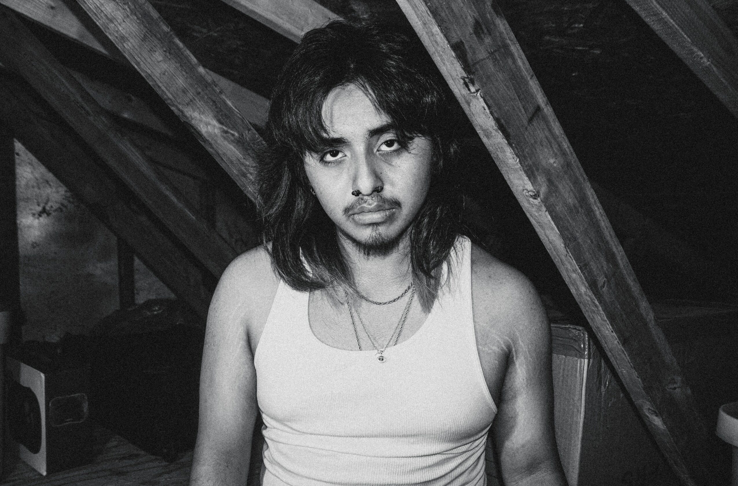 Young person with shoulder-length hair and a serious expression, wearing a sleeveless top, in a wooden attic space.