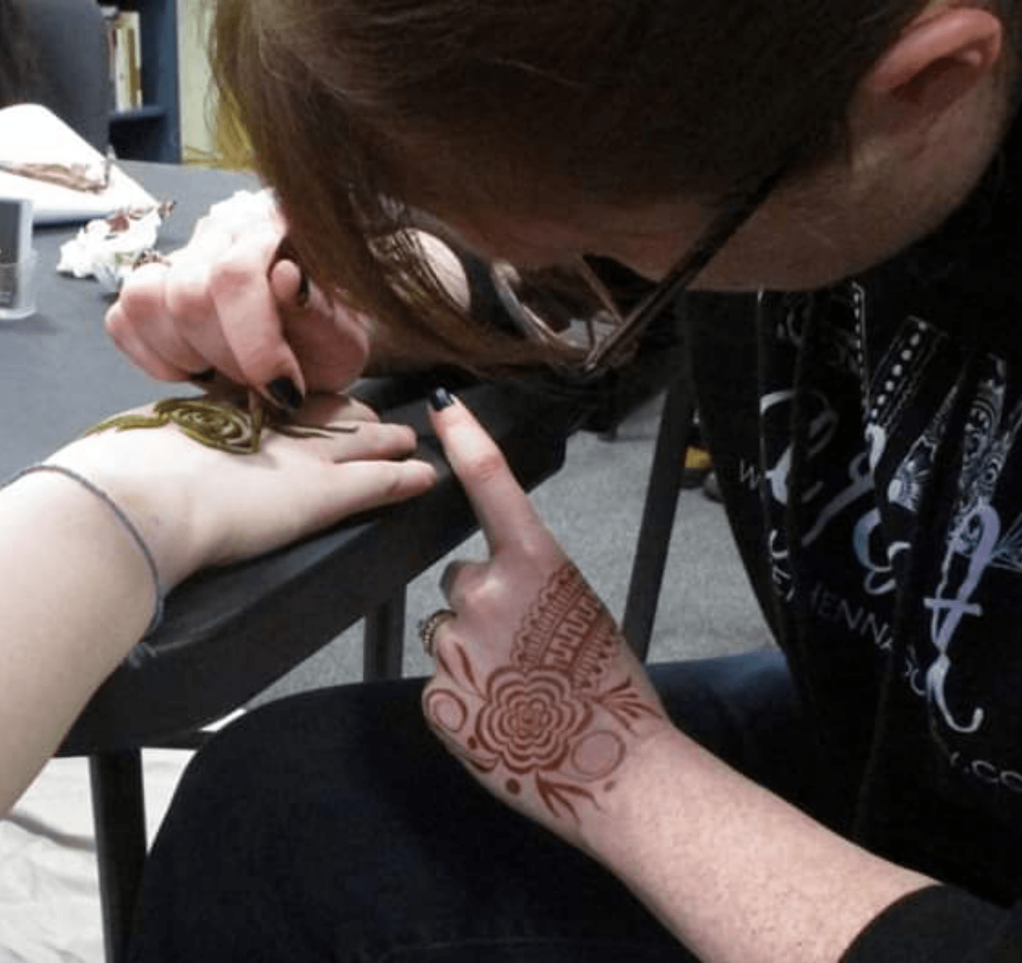Person getting a henna tattoo on hand, with artist applying design, in an indoor setting.